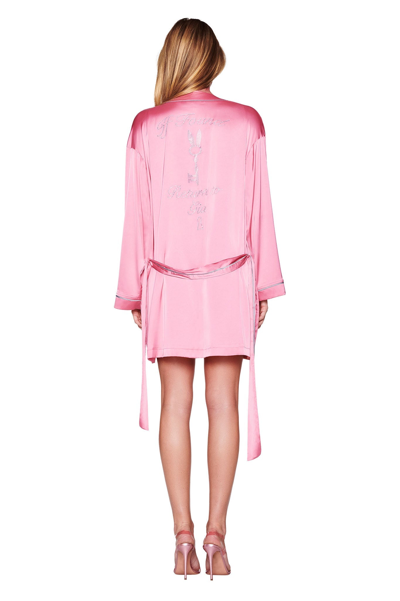 A person with long hair stands facing away, showcasing the relaxed fit of the I.AM.GIA MARTINA SLEEP ROBE in pink, with custom diamante 'GIA' text embroidered on the back. The satin robe is tied at the waist and is perfectly complemented by matching pink high-heeled shoes against a plain white background.
