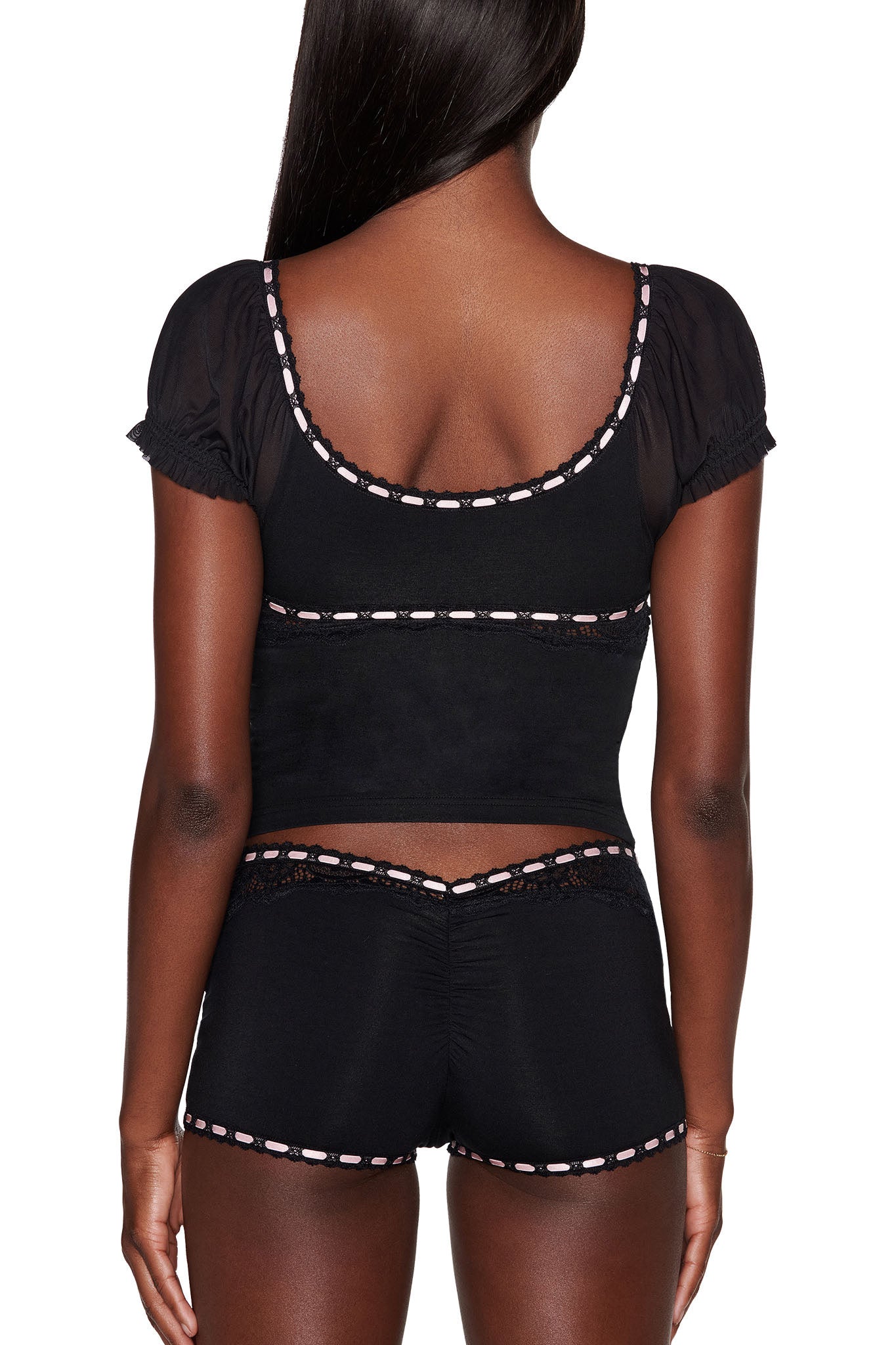 A person with long, dark hair is dressed in the I.AM.GIA DESTINY 2 Piece Set - Black, featuring a vintage-inspired short-sleeved top and matching shorts adorned with pink lace trim. They face away from the camera against a plain white background, highlighting the ensemble's bodycon fit.
