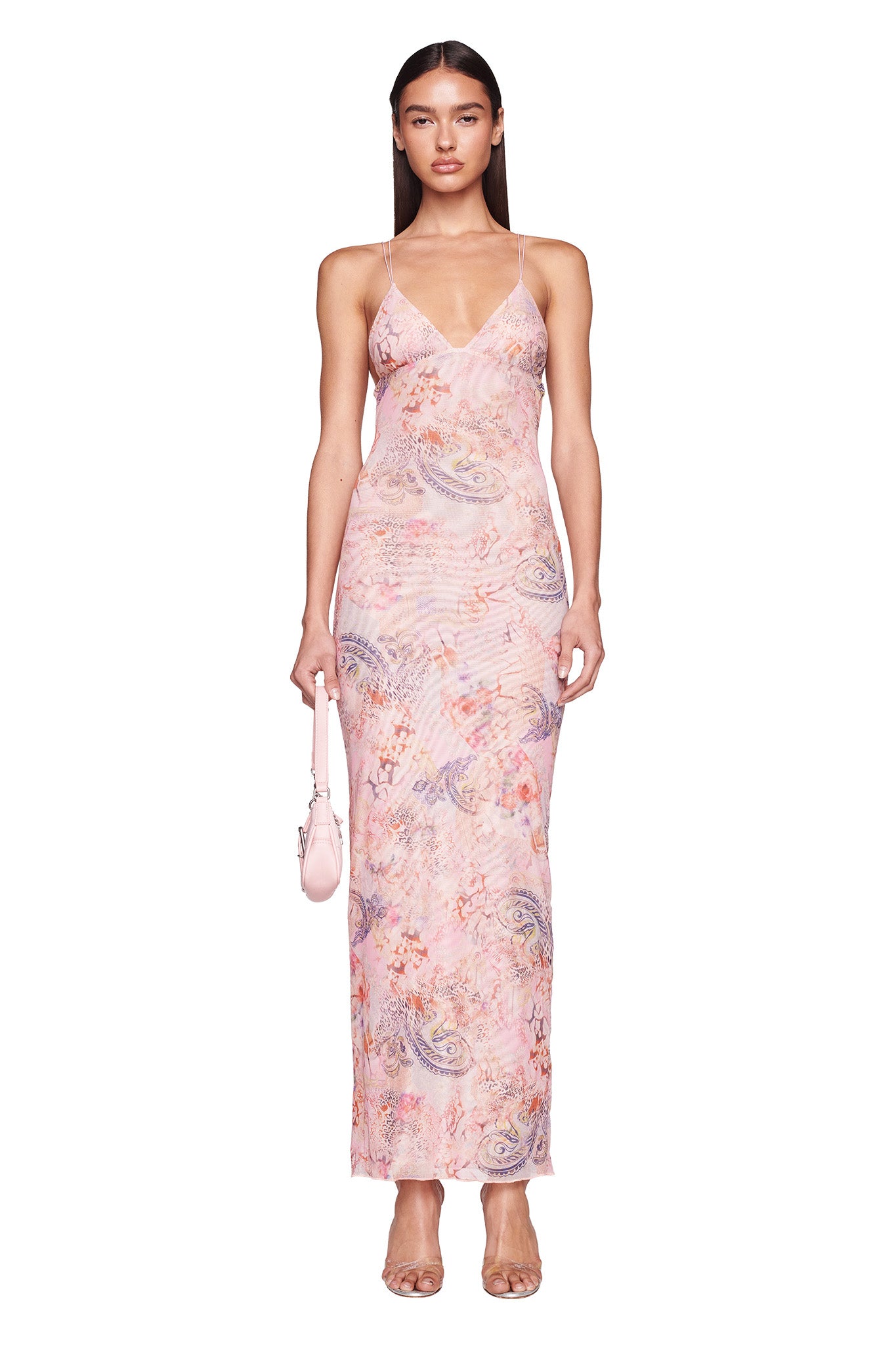 A person stands facing forward, wearing the I.AM.GIA ELLERY DRESS in pink with a floral and paisley pattern. They hold a small, light pink handbag and wear transparent high-heeled shoes. Their long, straight hair exudes I.AM.GIA vibes, while they maintain a serious expression.