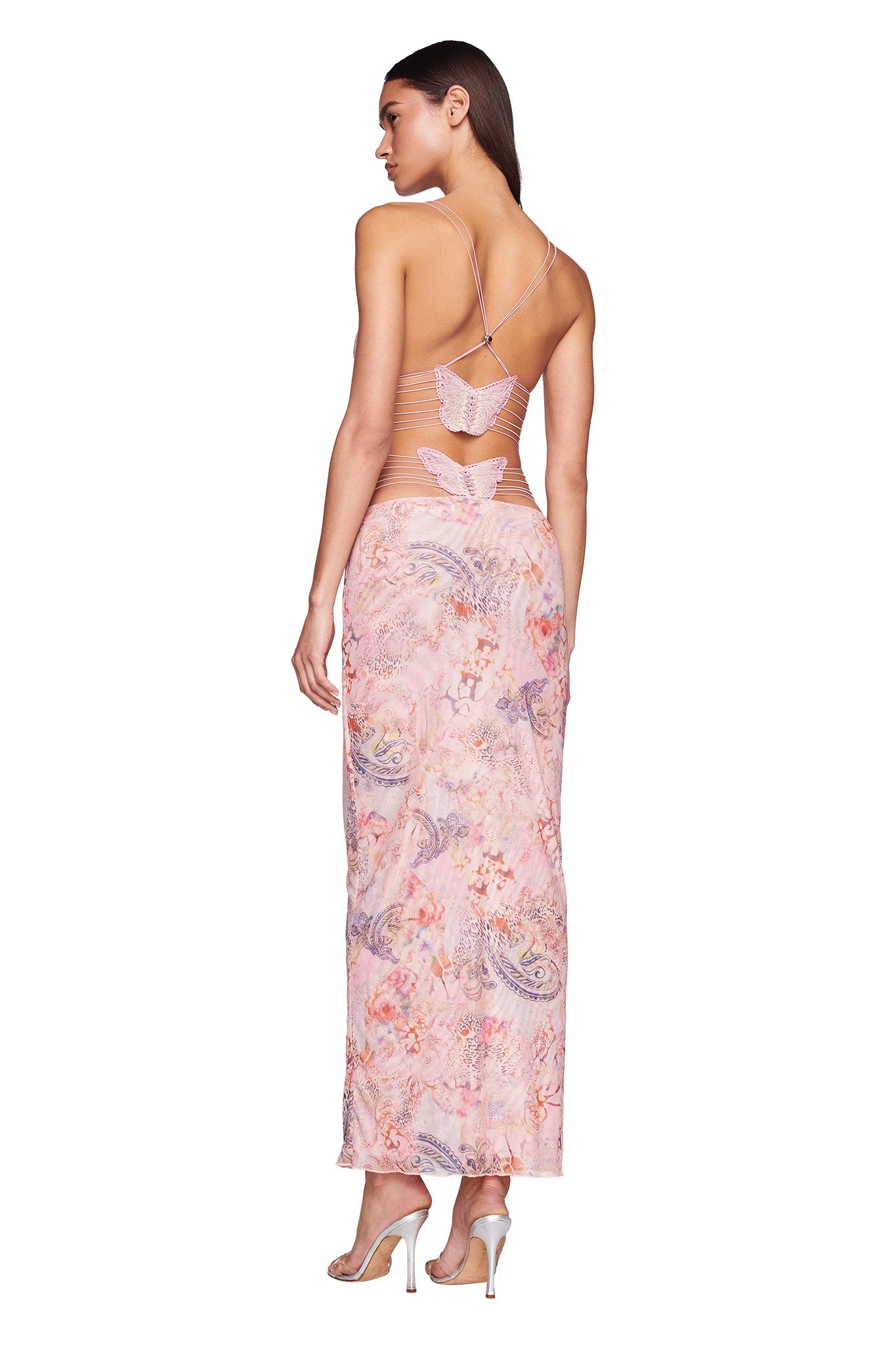 A person stands facing away, wearing the ELLERY DRESS - PINK : FLORA PAISLEY by I.AM.GIA. The pastel-colored floral and paisley dress features thin cross-back straps and a low-cut back with a butterfly-shaped design. The individual complements the look with high-heeled sandals and long hair flowing down.