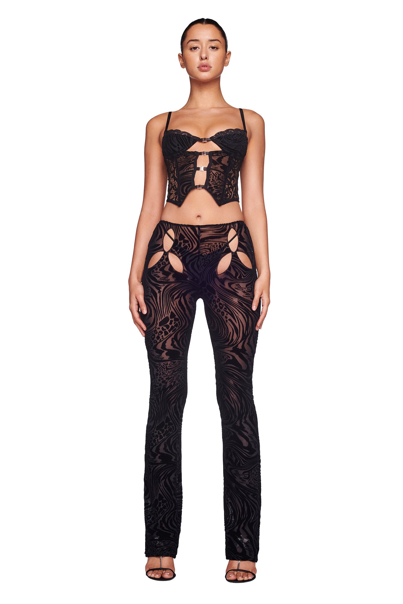 A woman is wearing an I.AM.GIA black lace outfit with intricate patterns. The top is a corset style with thin straps and cut-out details, and the matching AMINA PANT - BLACK BURNOUT has side cut-outs. She stands against a plain white background, looking straight ahead.