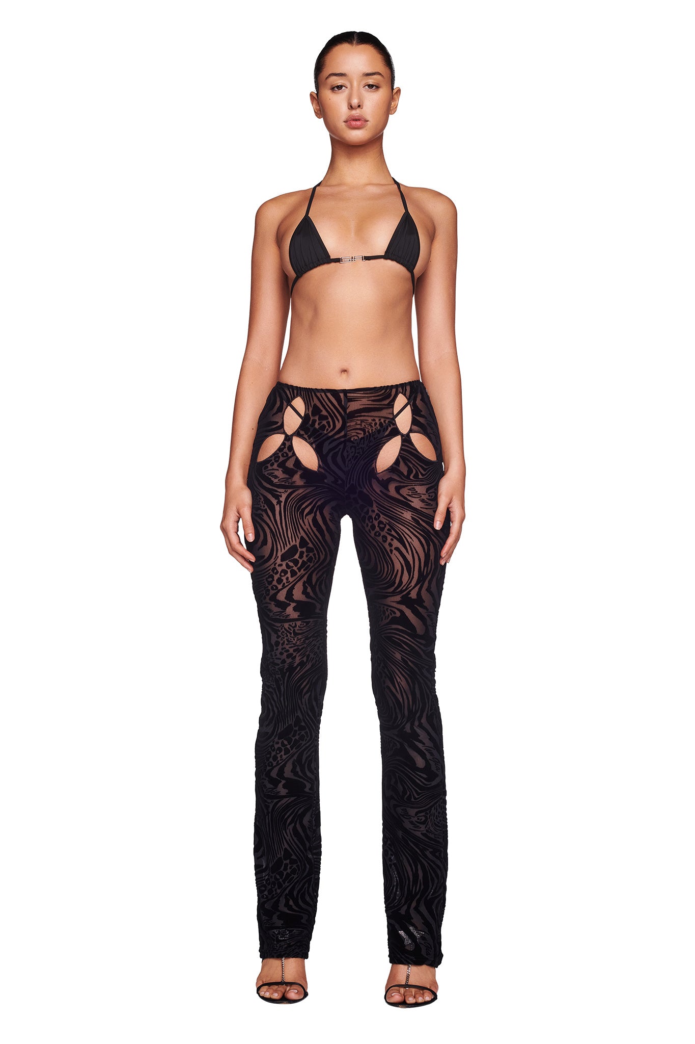 A woman stands against a plain white background, wearing the AMINA TOP - BLACK BURNOUT from I.AM.GIA. She pairs it with high-waisted, sheer black pants that feature a swirling pattern and cutout detailing at the hips. Her hair is pulled back and she's wearing black high-heeled sandals. She is facing forward.