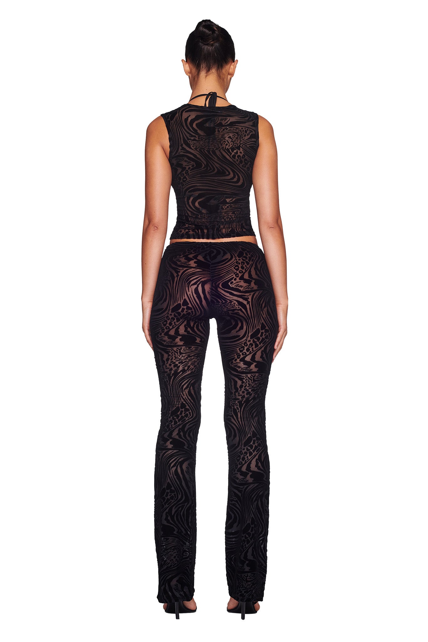 A woman is standing facing away from the camera, fully outfitted in I.AM.GIA. She wears a sleeveless, form-fitting black top and the AMINA PANT - BLACK BURNOUT, which features a swirling, sheer pattern. Her hair is styled in a high bun, and she completes her look with black high heels against a white background.