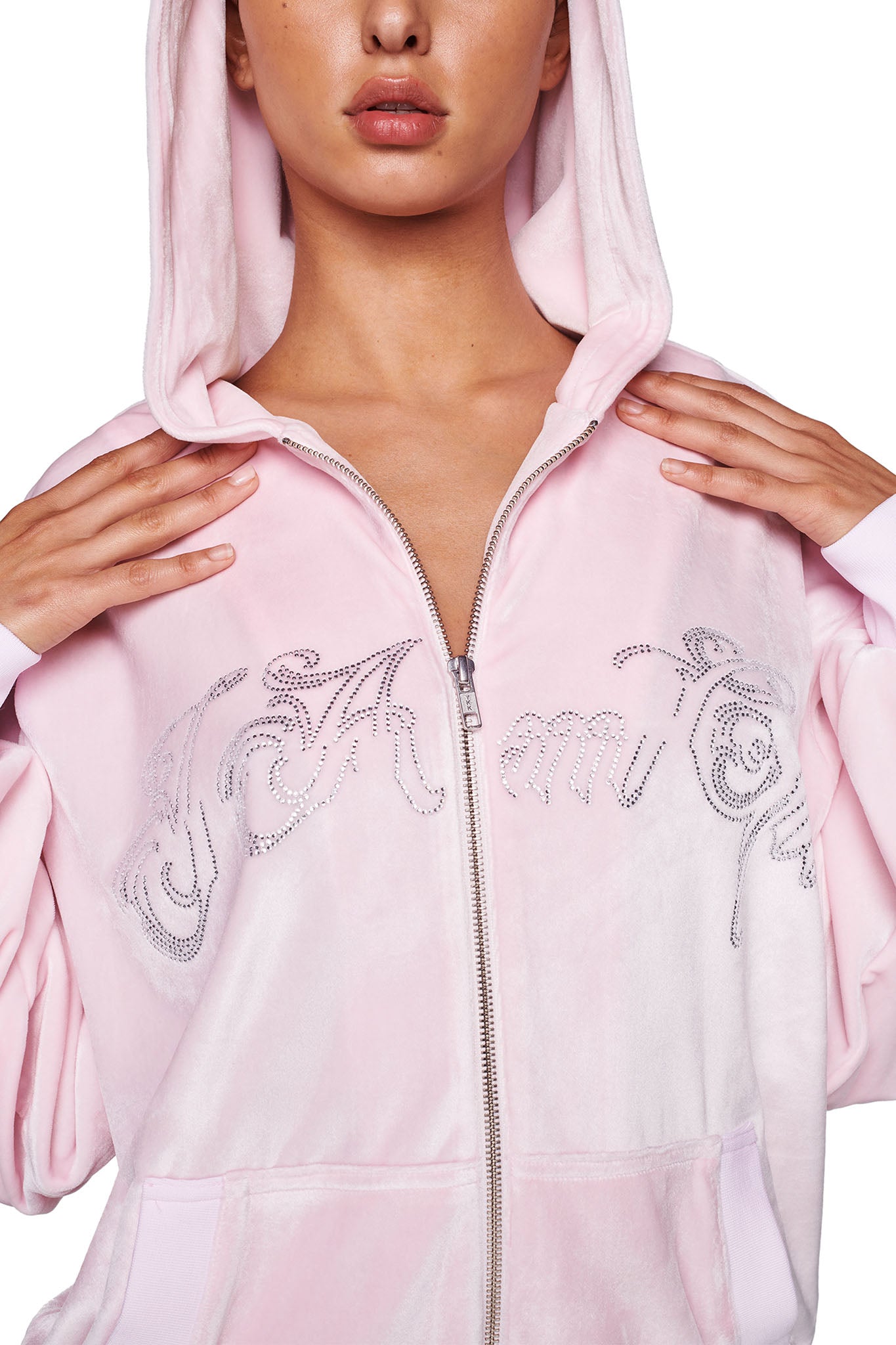 A person wearing an I.AM.GIA DARIA HOODIE in Icy Pink with the hood up and partially zippered down. The hoodie features silver rhinestone embellishments forming a design or text on the chest area. The background is plain white.