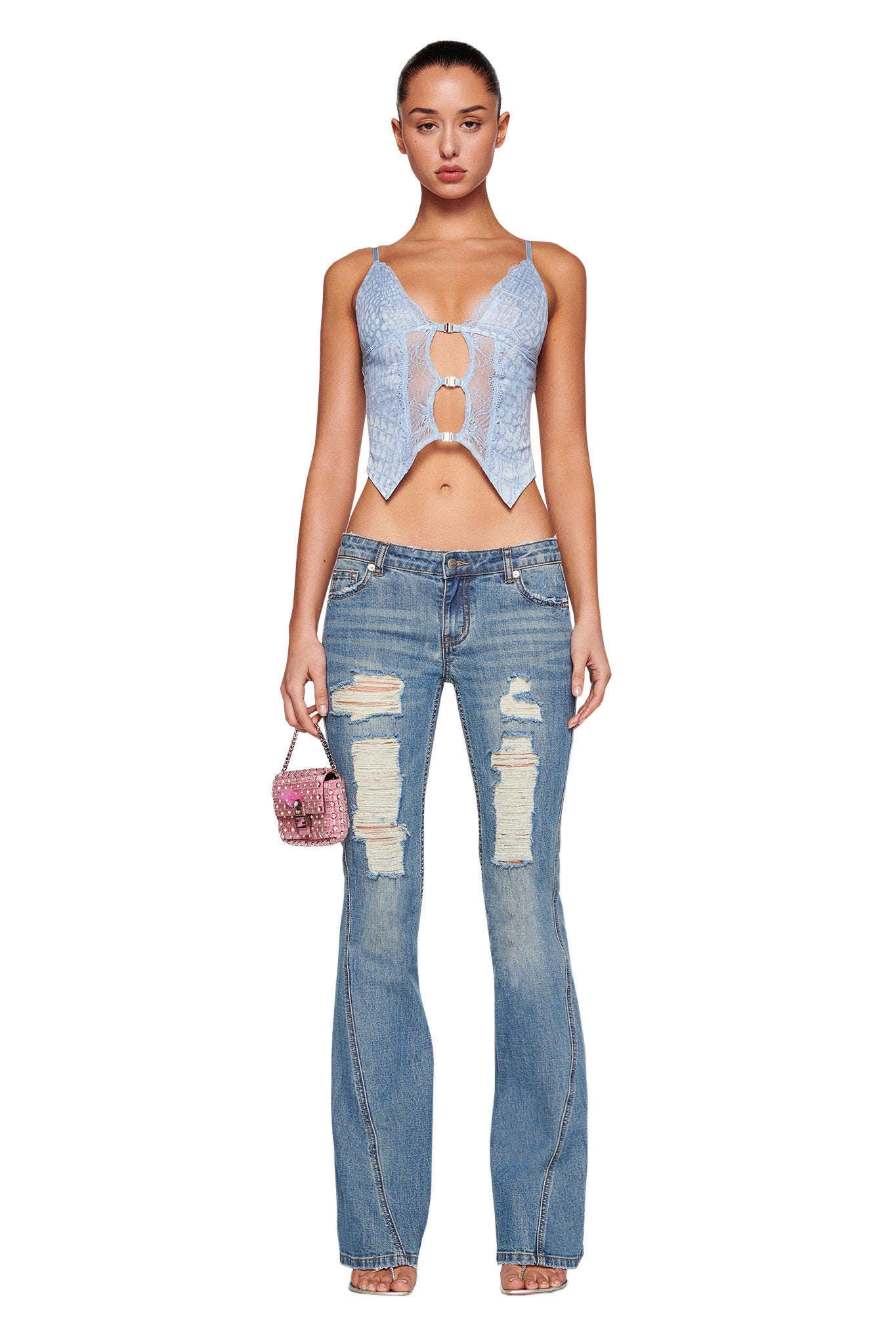A woman is standing against a white background, wearing an I.AM.GIA light blue lace-up crop top and MYLEE JEAN - BLUE : DENIM BLUE from I.AM.GIA. She holds a small pink purse in her left hand and is wearing clear high-heeled sandals. Her hair is pulled back, and she has a neutral expression.