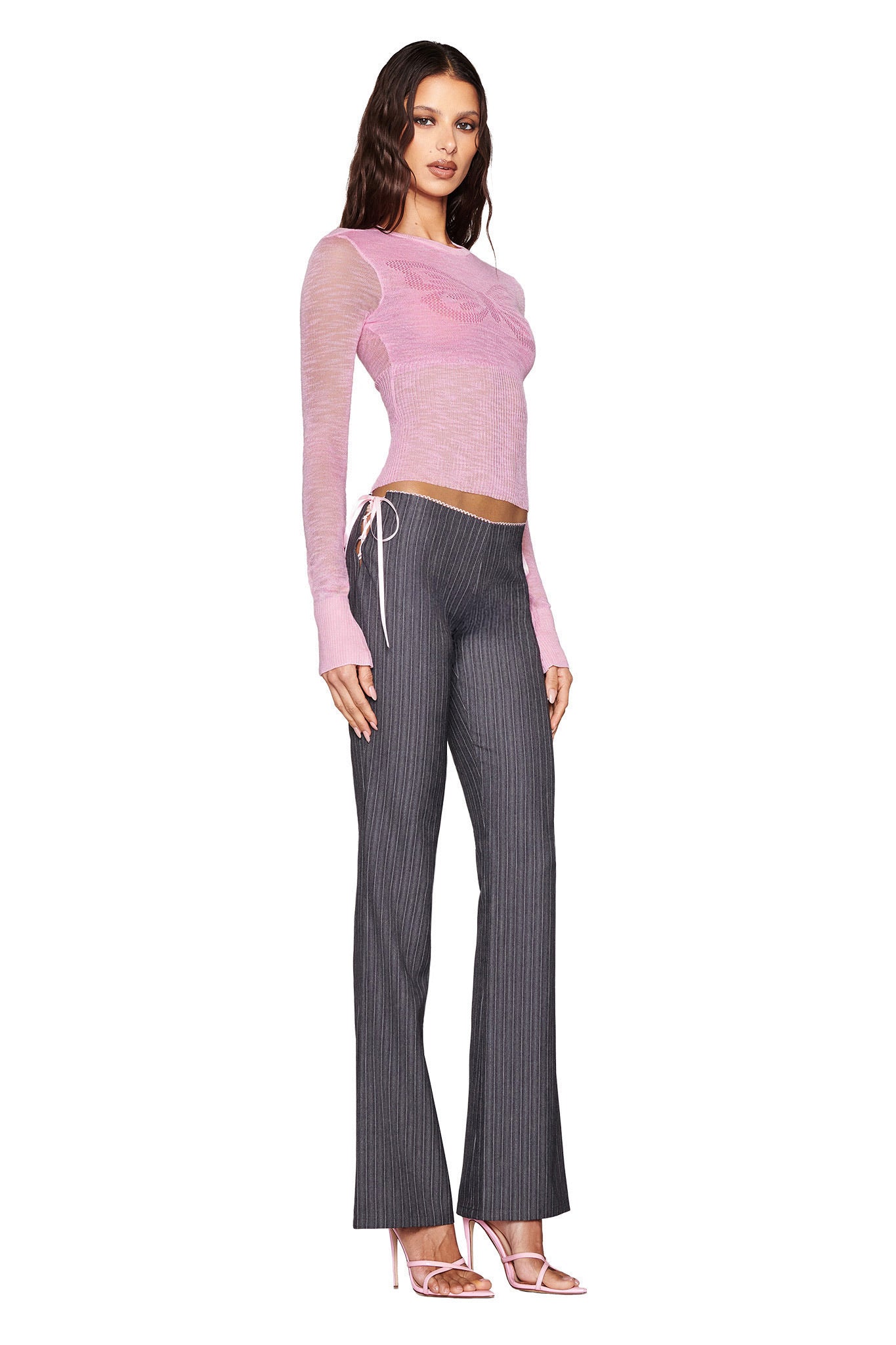 A woman poses against a white background wearing the I.AM.GIA MARIPOSA Top in an ultra-light sheer pink with gray pinstripe pants. She completes her chic look with long brown hair, pink high-heeled sandals, and a ribbed hem for added elegance.