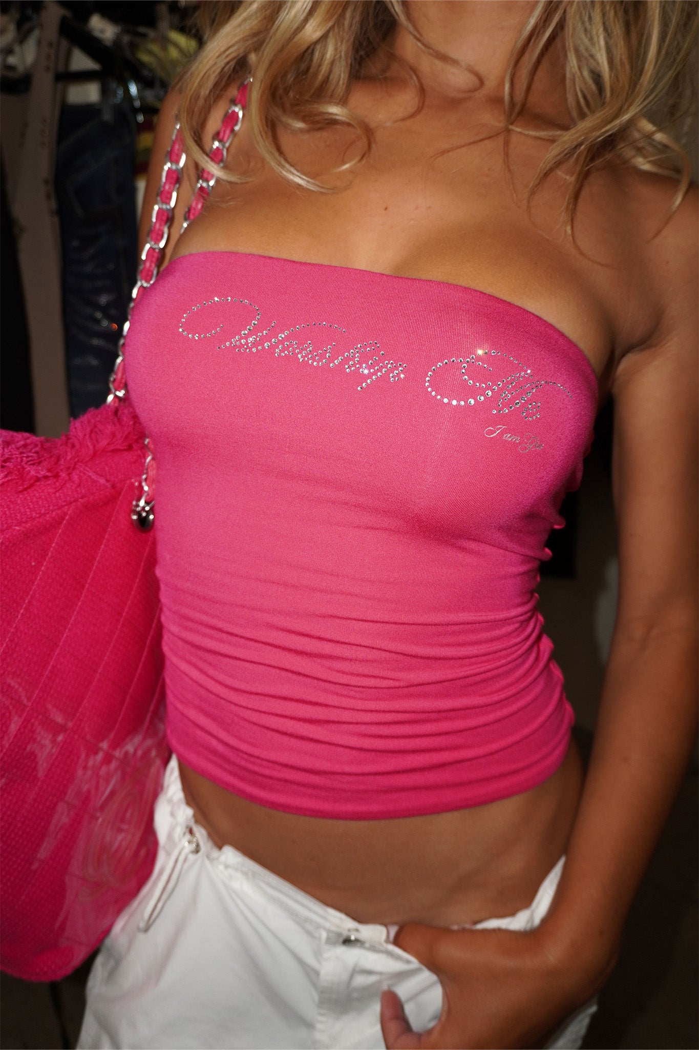 A woman wears the I.AM.GIA NESSY BOOB TUBE - WORSHIP ME in hot pink with a rhinestone graphic, paired with white shorts and a large pink bag. Her blonde hair is partially visible, and the background is blurred.