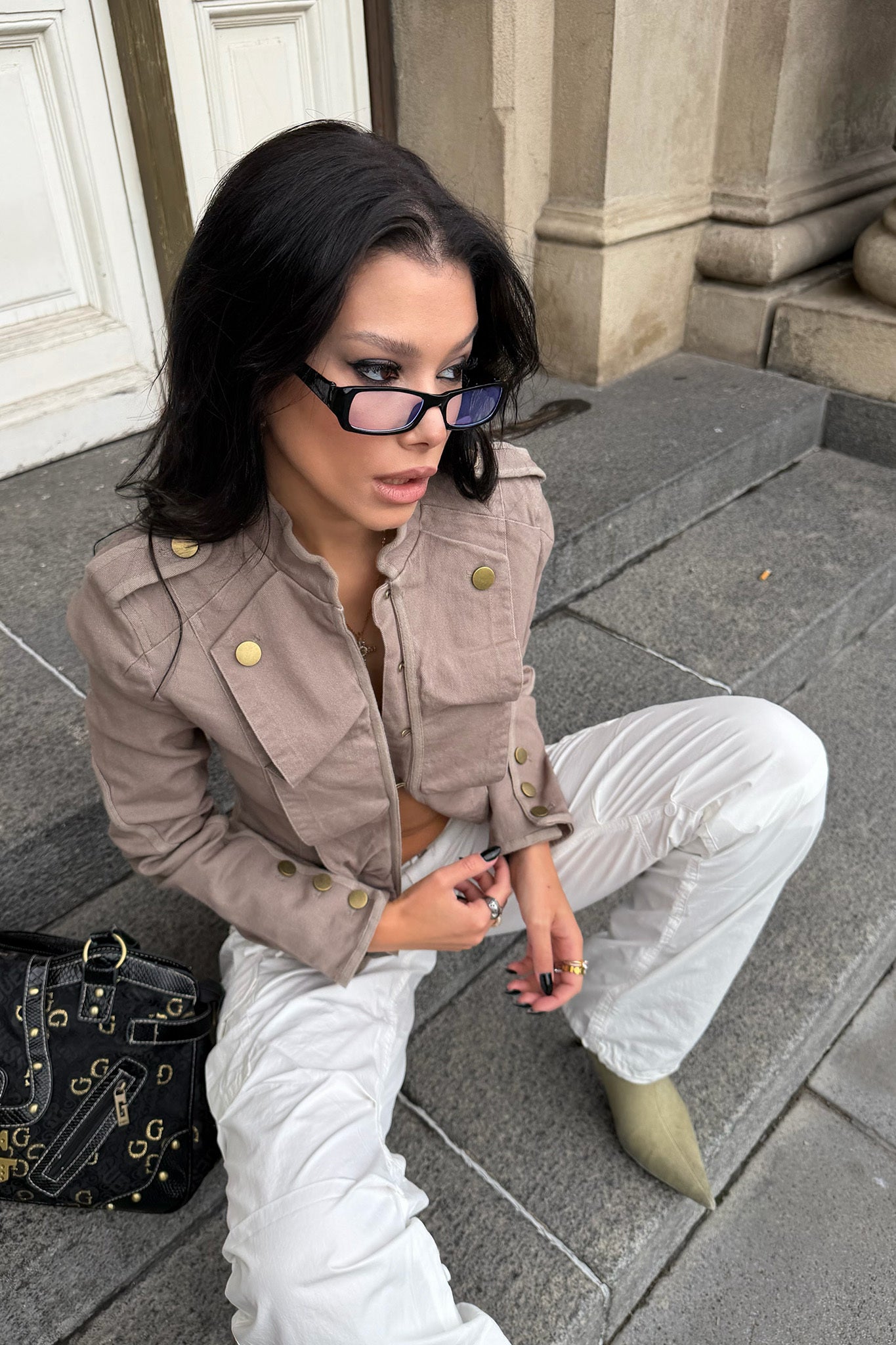 A woman with long dark hair, wearing the I.AM.GIA SHARN JACKET in Mushroom, white pants, and glasses, sits on stone steps by a building entrance. She holds a cigarette and has a bag beside her.