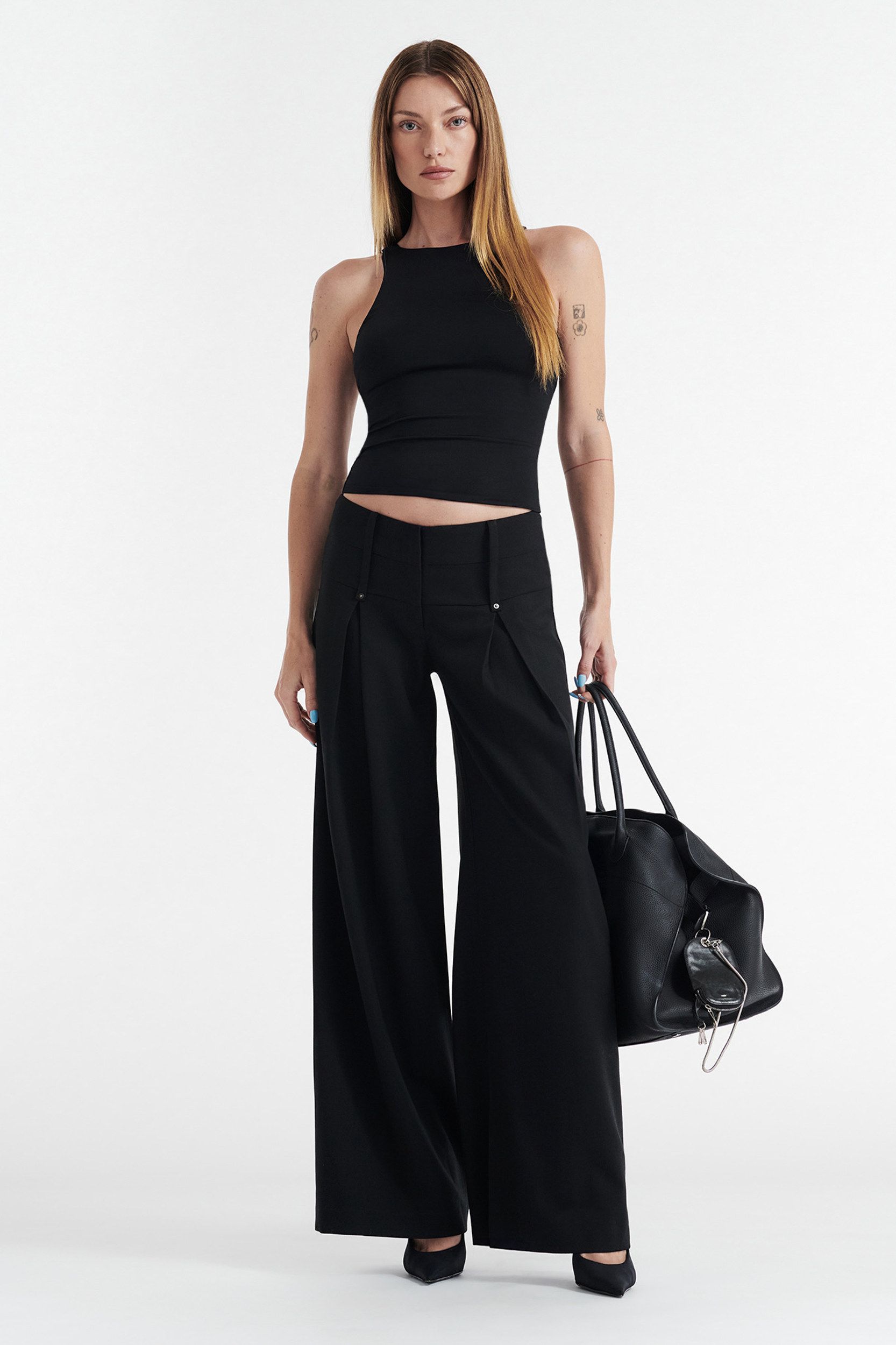 A woman with straight, long hair wears the CDP SOPHIE BASIC SLEEVELESS TANK WITH SHELF BRA in black, paired with high-waisted wide-leg black pants and heels. She holds a large black handbag, standing against a white background.