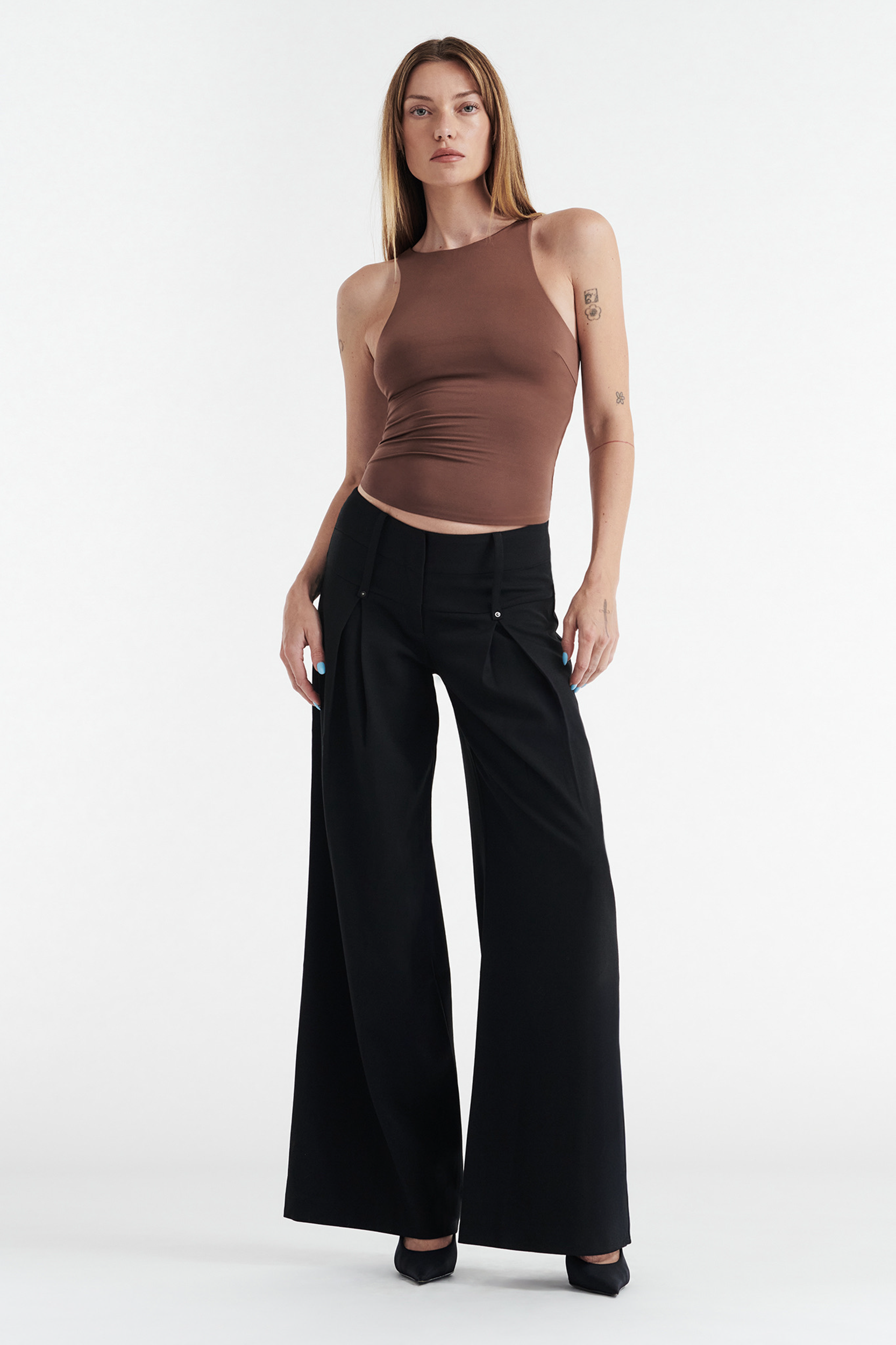 A woman stands against a white background in the CDP SOPHIE BASIC SLEEVELESS TANK WITH SHELF BRA in chocolate, paired with high-waisted wide-leg black pants and black heels. Her straight hair is down, and she has a relaxed, poised look.