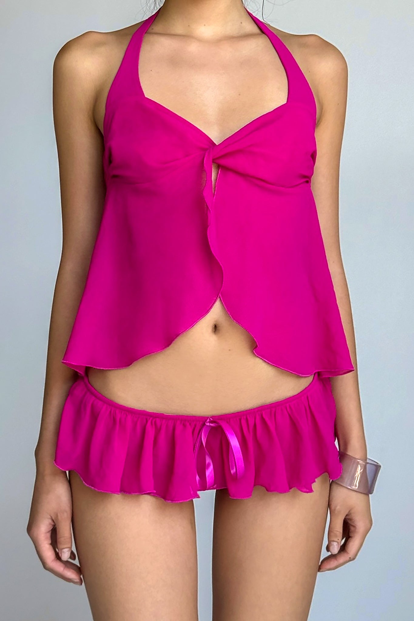 A person is wearing the CHEYENNE SET - PINK from I.AM.GIA, a matching hot pink sleep set in georgette fabric. The outfit consists of a semi-sheer sleeveless top with a bow at the chest and frilly, ruffled shorts. Their left hand is resting by their side, adorned with a clear bracelet. The background is plain and neutral.