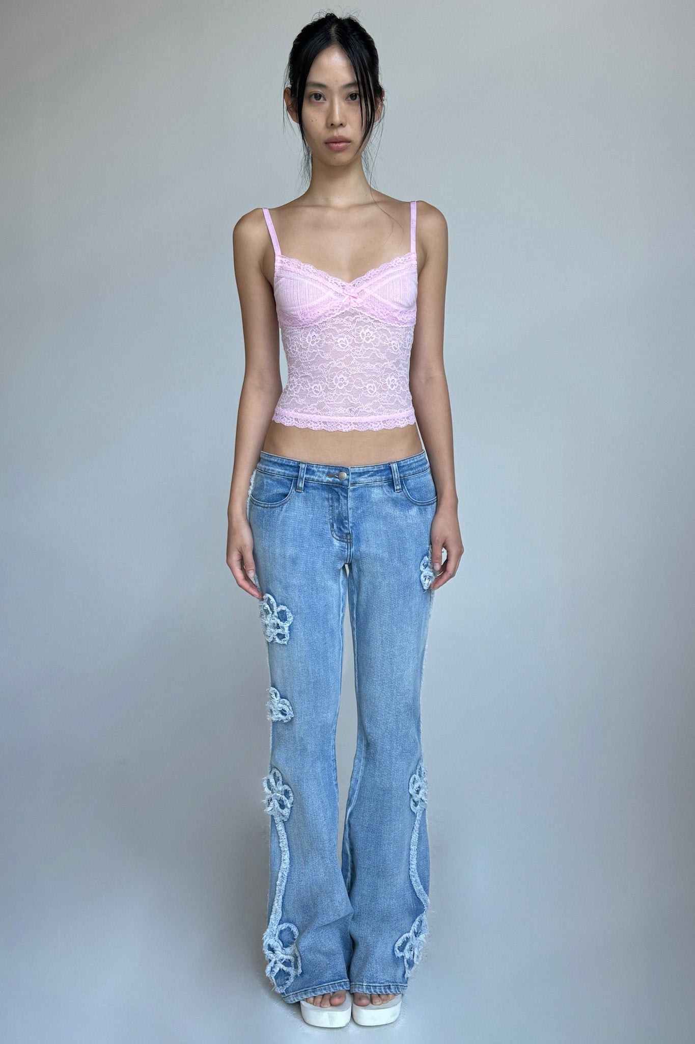 A woman stands against a plain background, wearing the SERINA TANK - PINK : LACE camisole from I.AM.GIA and light blue jeans with floral embroidery on the legs. She has long dark hair tied back and is looking directly at the camera. She is also wearing white shoes.