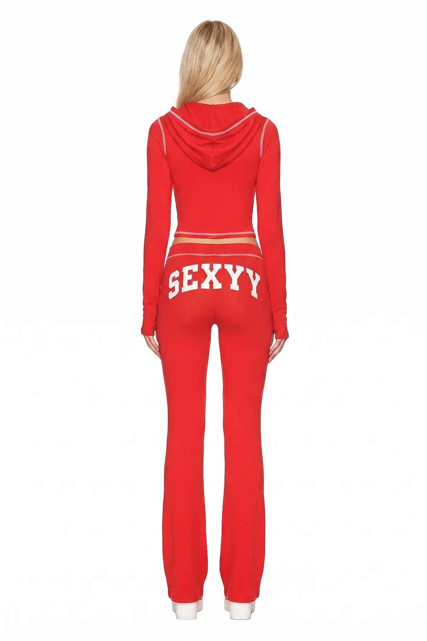 A person with long blonde hair is seen from behind wearing the I.AM.GIA X SEXYY RED BLARE HOODIE by I.AM.GIA, a red hooded tracksuit featuring bold white "SEXY Y" lettering across the back of the pants and white stripes.