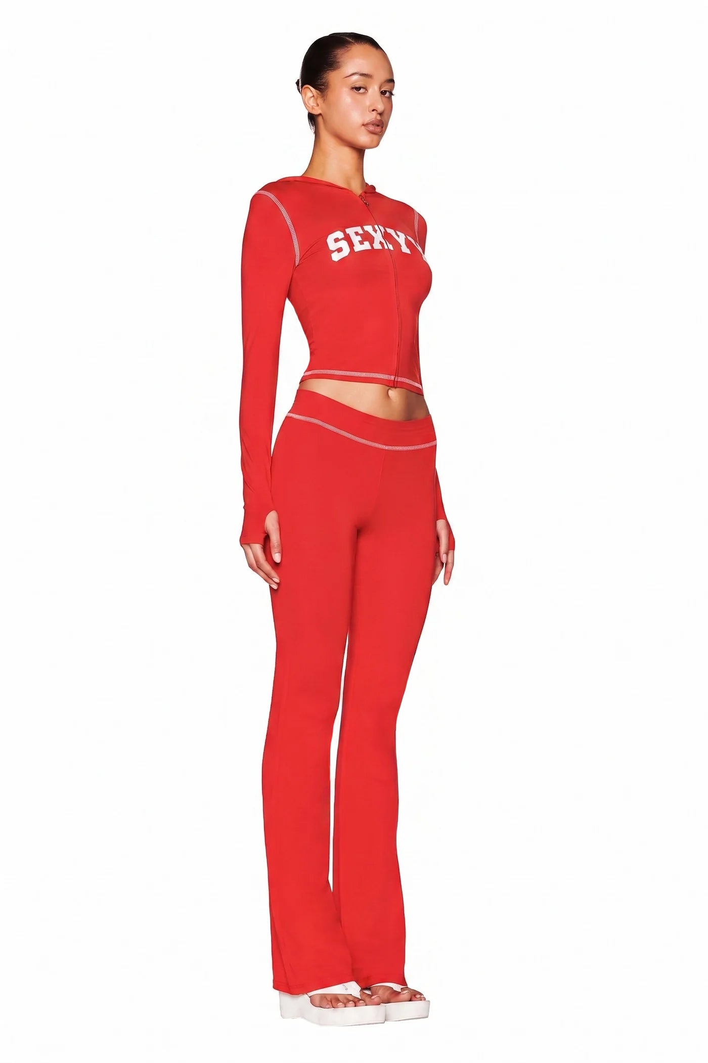 A woman stands in profile in a fitted, bright red I.AM.GIA X SEXYY RED BLARE TRACKPANT set with a low-rise waist and long sleeves. The top features "SEX" in white letters and is paired with white platform sandals against a plain white background.