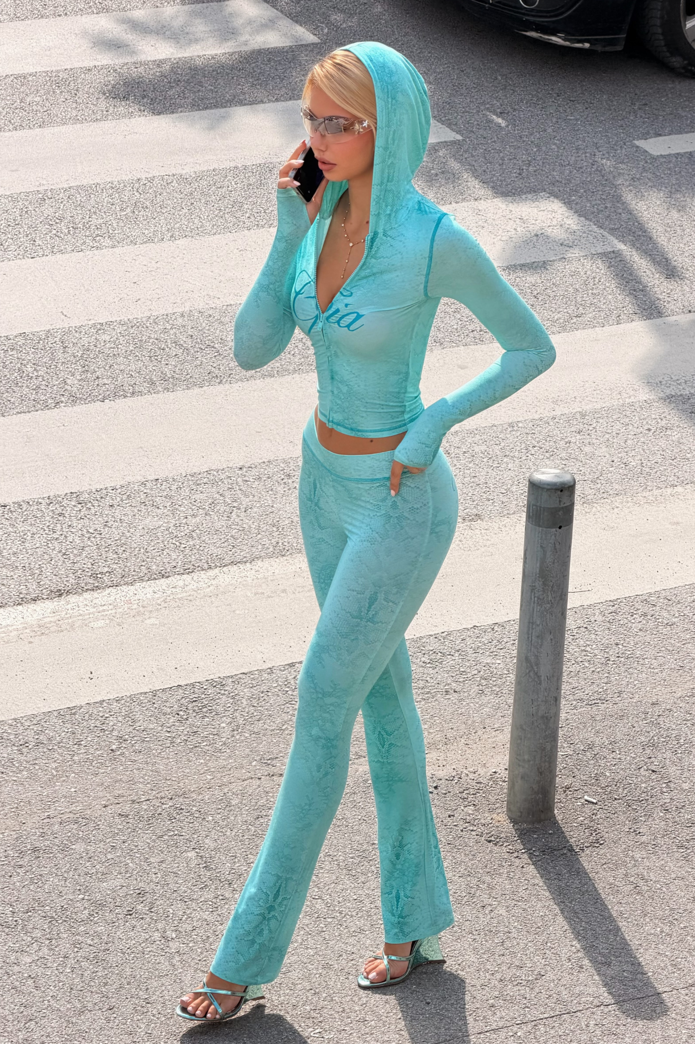 A woman, dressed in a light blue hooded ensemble and chic heeled sandals, walks along a city street while chatting on her phone. Sporting sunglasses as she crosses the crosswalk, her flared-leg pants—the BLARE TRACKPANT - TEAL SERPENTINE by I.AM.GIA—bring a trendy edge to her modern style.