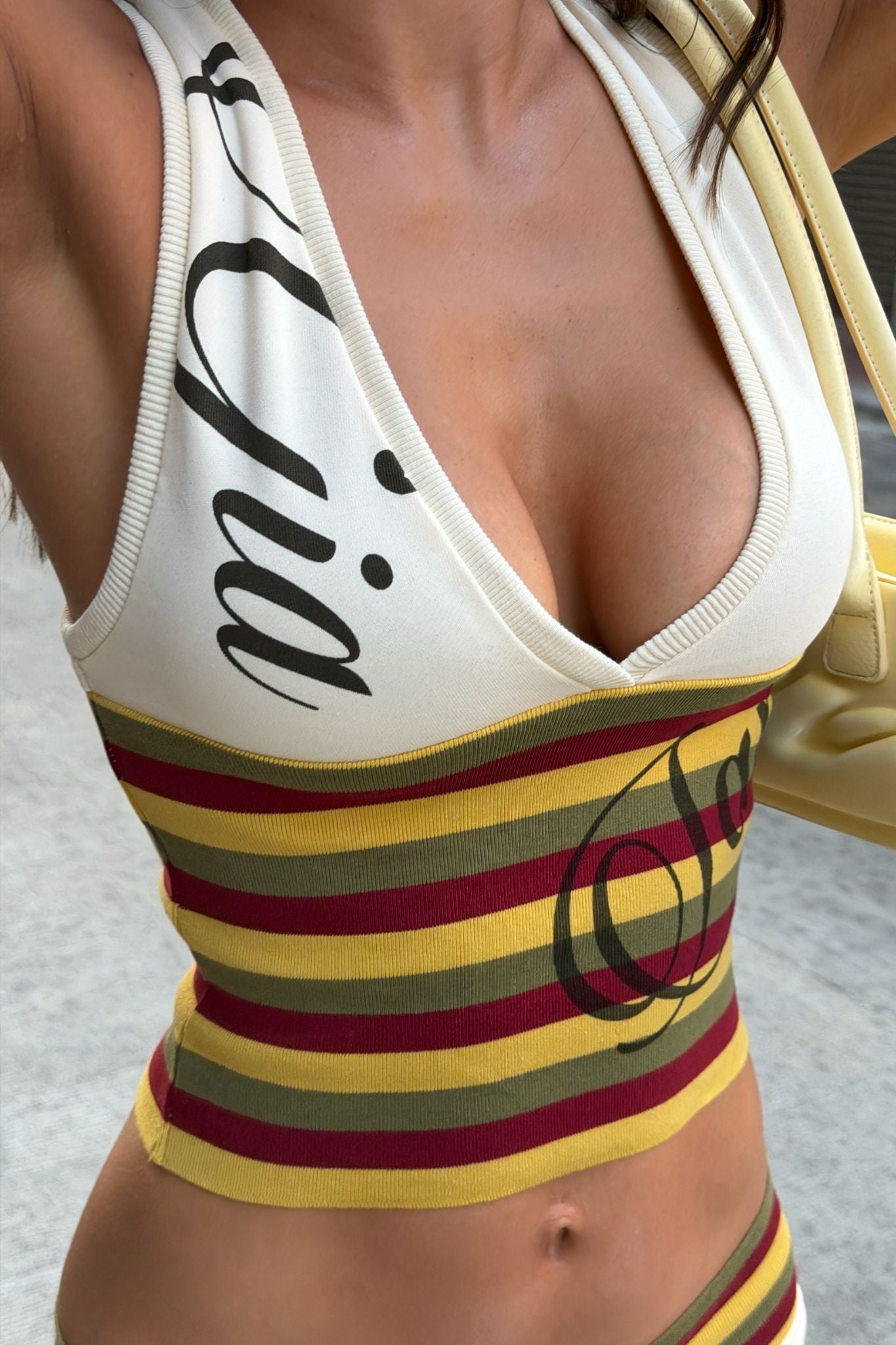 A person wears the I.AM.GIA CANDI TOP - CREAM, a bodycon V-neck with horizontal yellow, red, and brown stripes and cursive writing, paired with a light yellow handbag.