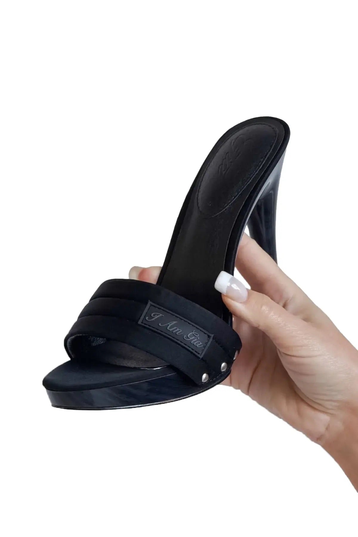 A manicured hand holds the I.AM.GIA NICKY HEEL in black neoprene, showcasing its sculpted stiletto heel, open toe, and metal nameplate on the padded vamp strap, set against a white background.