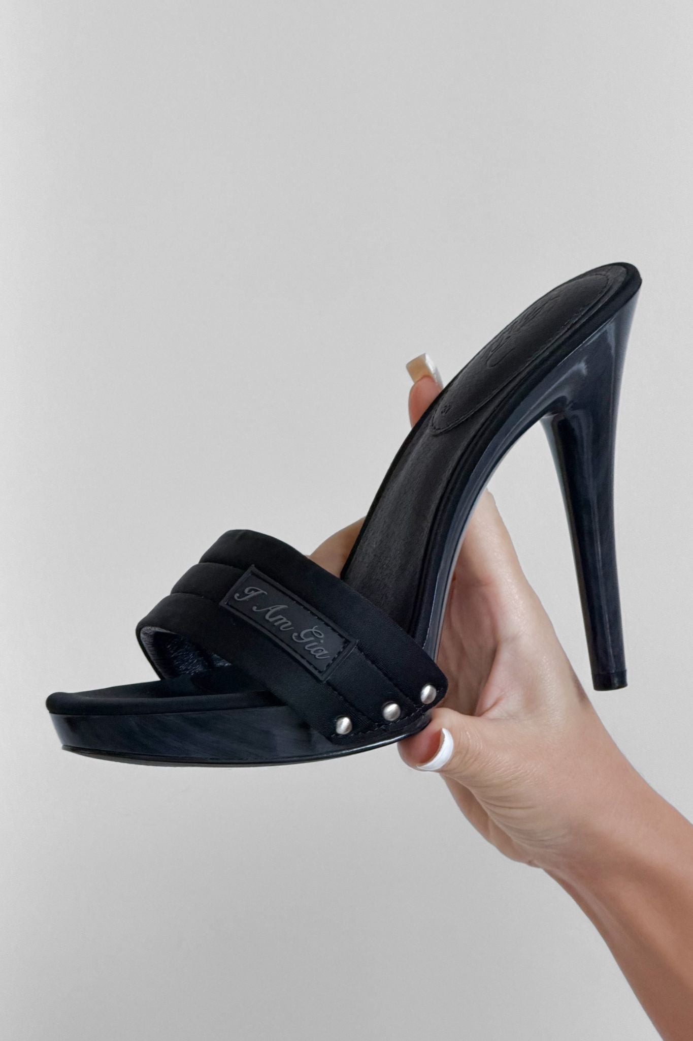 A hand holds up the I.AM.GIA NICKY HEEL in black neoprene, a platform sandal with a stiletto heel, wide padded vamp strap, and silver rivet accents against a plain light background.