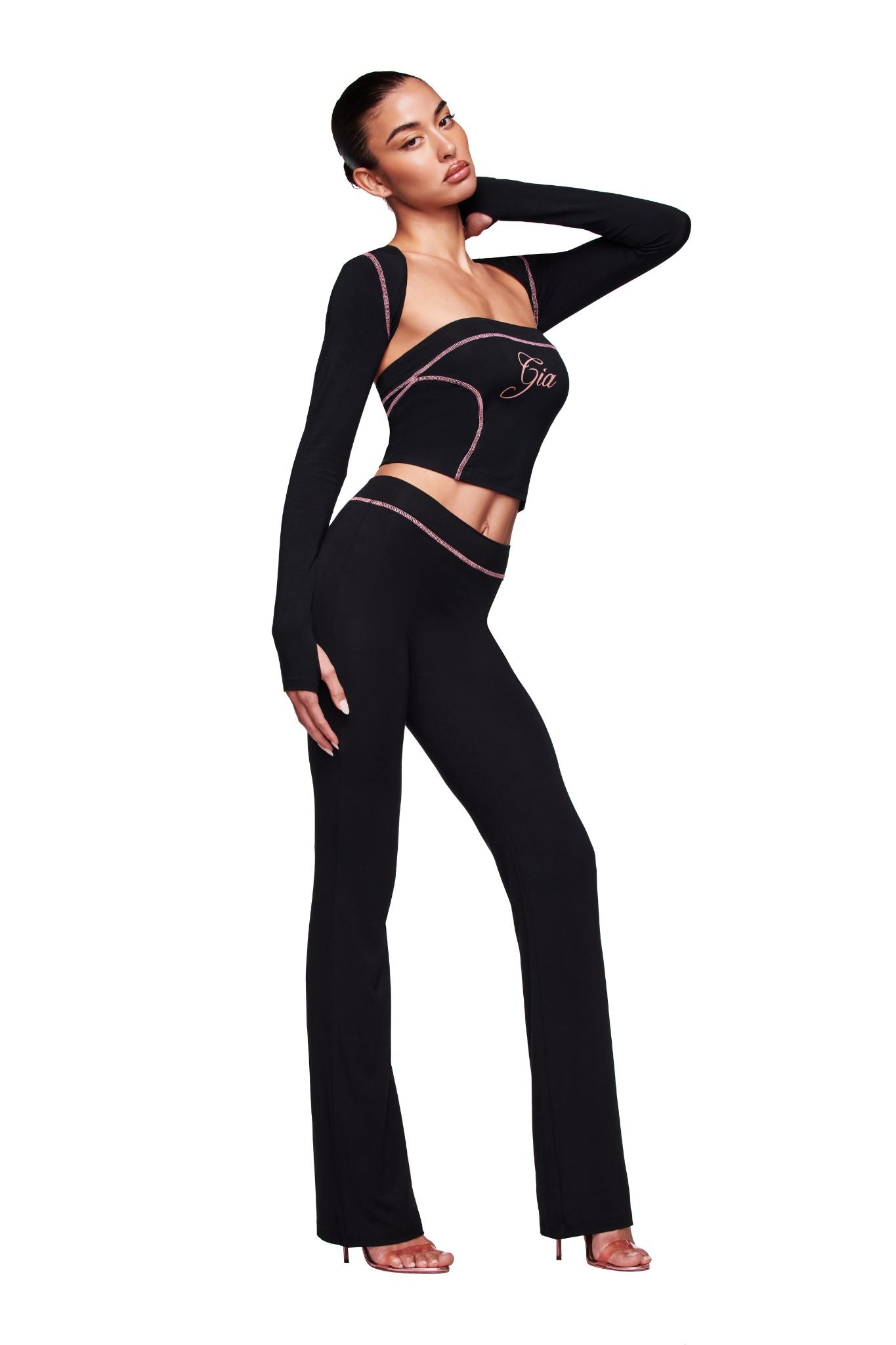A woman poses in the I.AM.GIA BLARE SHRUG - BLACK, a form-fitting black two-piece with pink trim. The long-sleeve top features a cut-out design and "Gia" on the chest. She stands in heels against a white background.