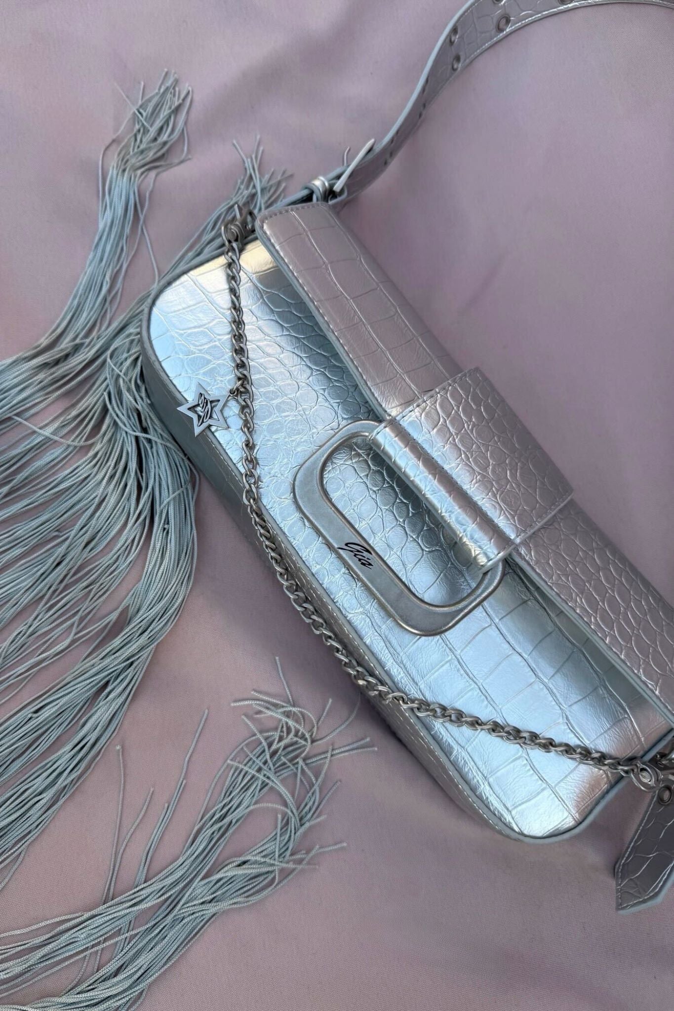 The MILA METALLIC BAG - SILVER METALLIC by I.AM.GIA, a mini shoulder purse with faux crocodile texture, chain strap, and long fringe accents, is displayed on a soft pink background with subtle I.AM.GIA branding.
