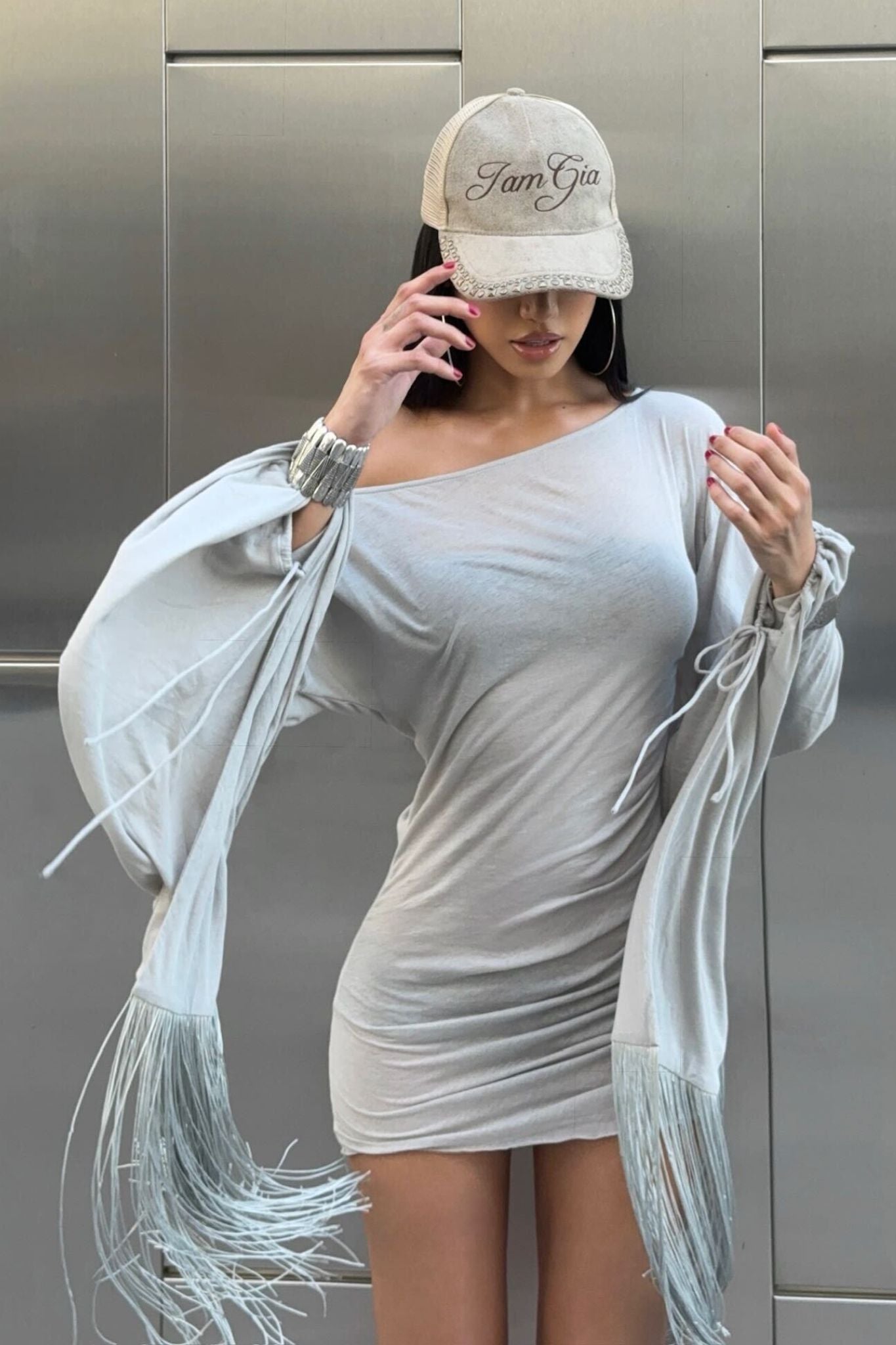 A woman in the I.AM.GIA LUMA MINI DRESS - GREY, paired with a matching cap, talks on the phone. She stands against a metallic wall, her face partially hidden by the cap.