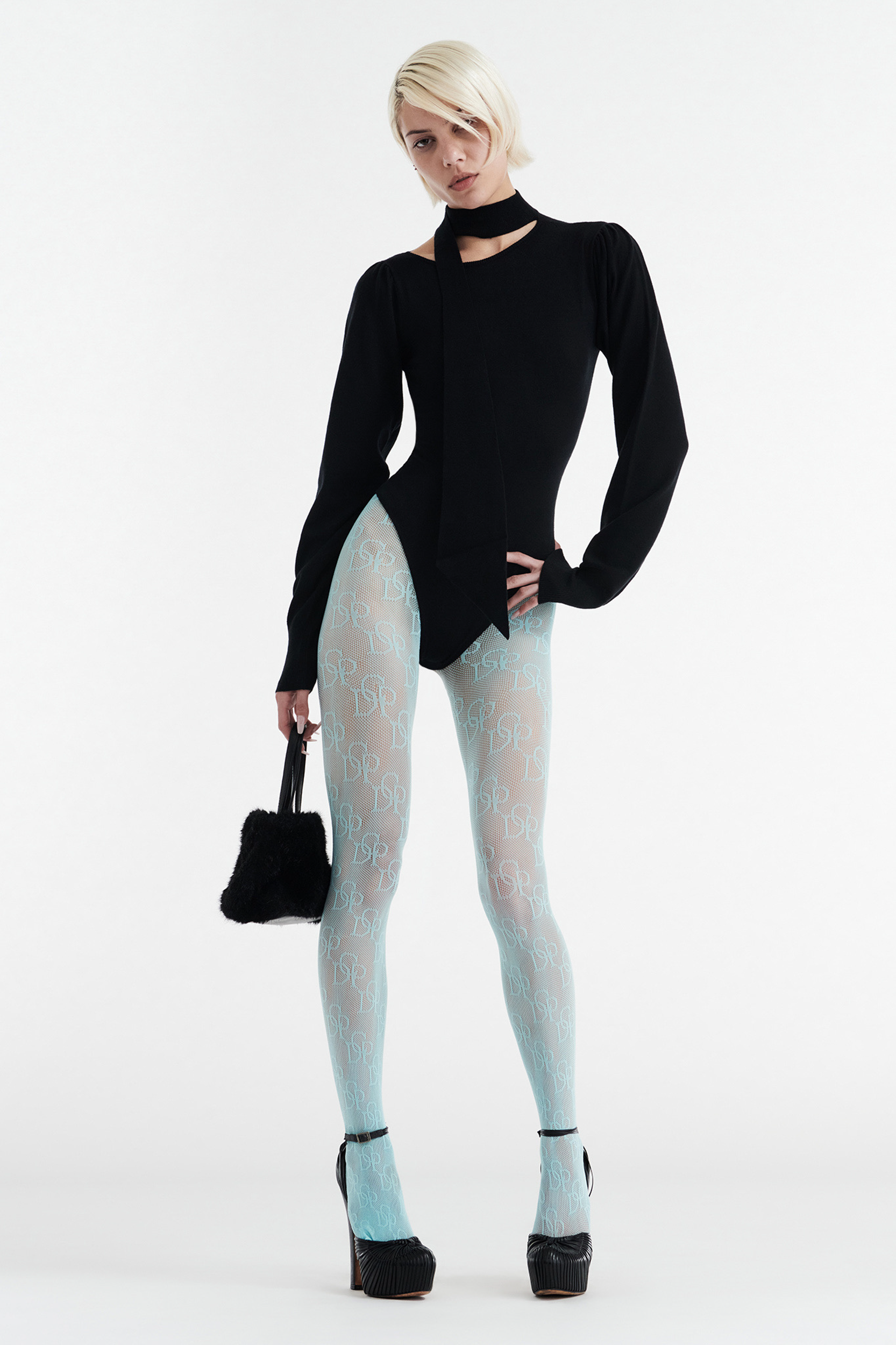 A person with short blond hair poses in the CDP VIVIENNE LONG SLEEVE KNIT BODYSUIT in black, paired with a necktie detail, sheer light blue patterned tights, black platform heels, and a small black handbag against a white background.