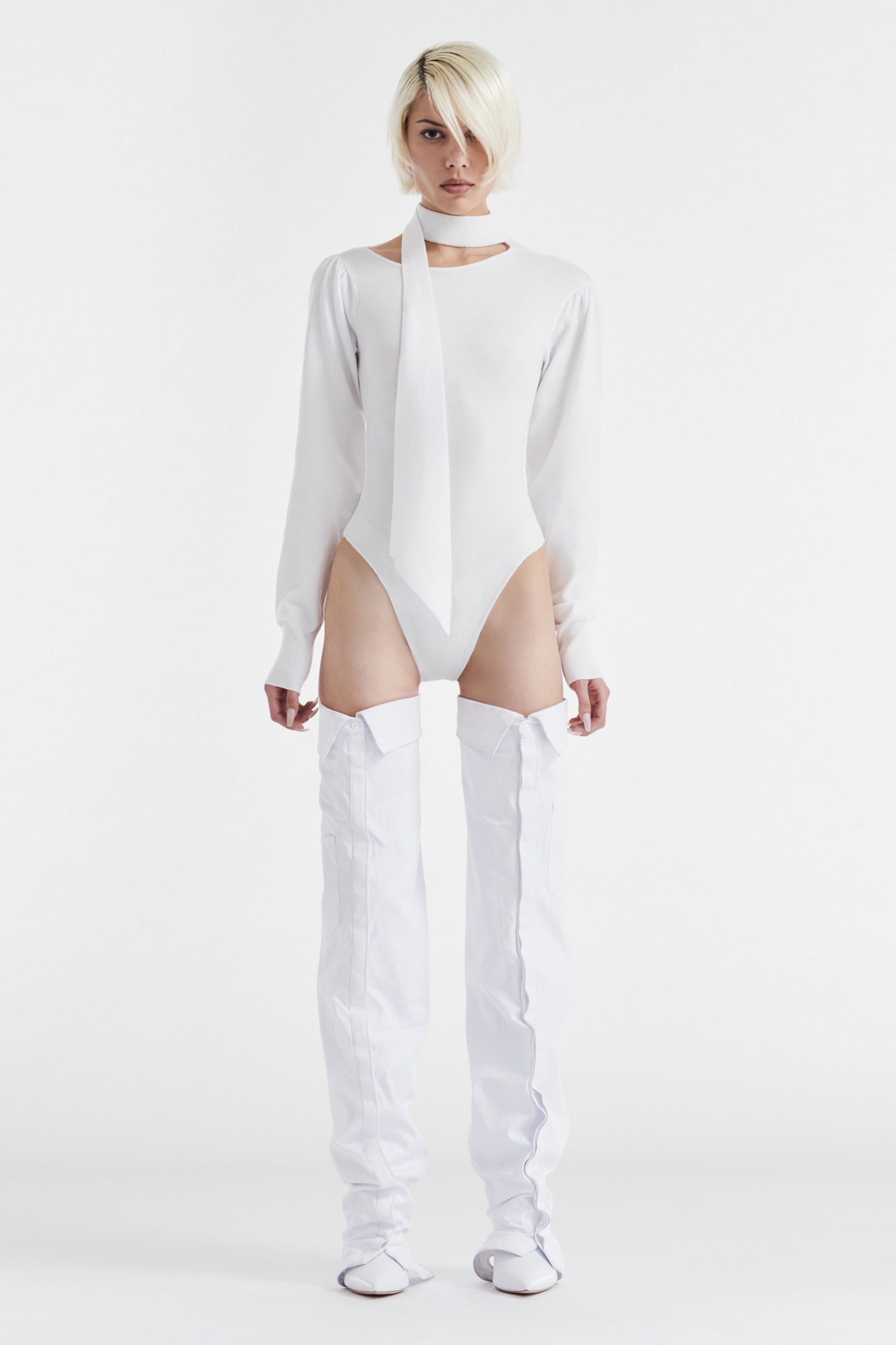 A person with short blond hair models the CDP VIVIENNE LONG SLEEVE KNIT BODYSUIT in white, paired with a matching necktie and thigh-high white boots, against a plain white background.