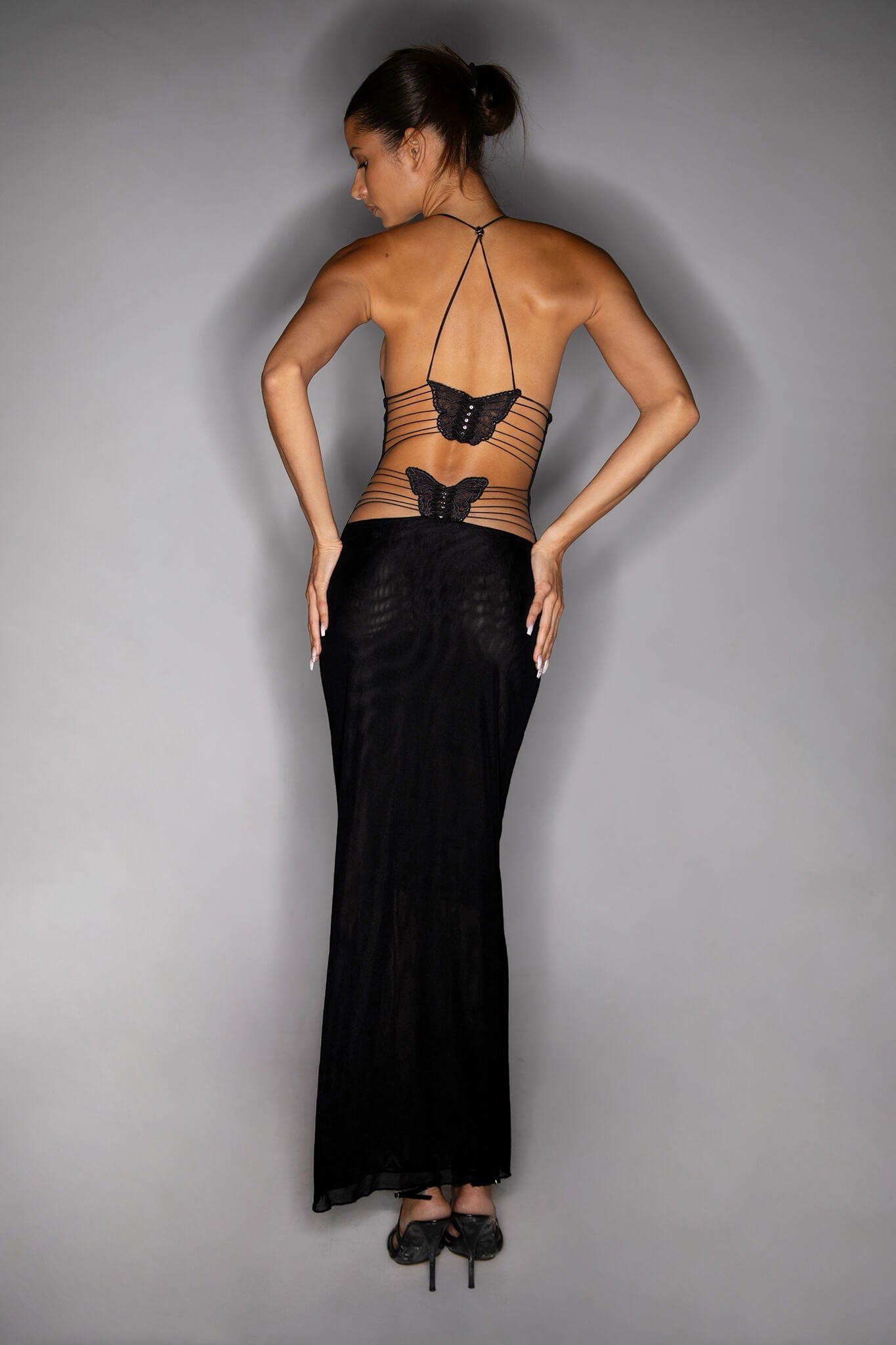 A woman stands facing away, showcasing the ELLERY DRESS - BLACK by I.AM.GIA. The ankle-length dress features an open back with butterfly accents at the mid-back and thin straps. Her hair is tied in a bun, and she is wearing black high heels.
