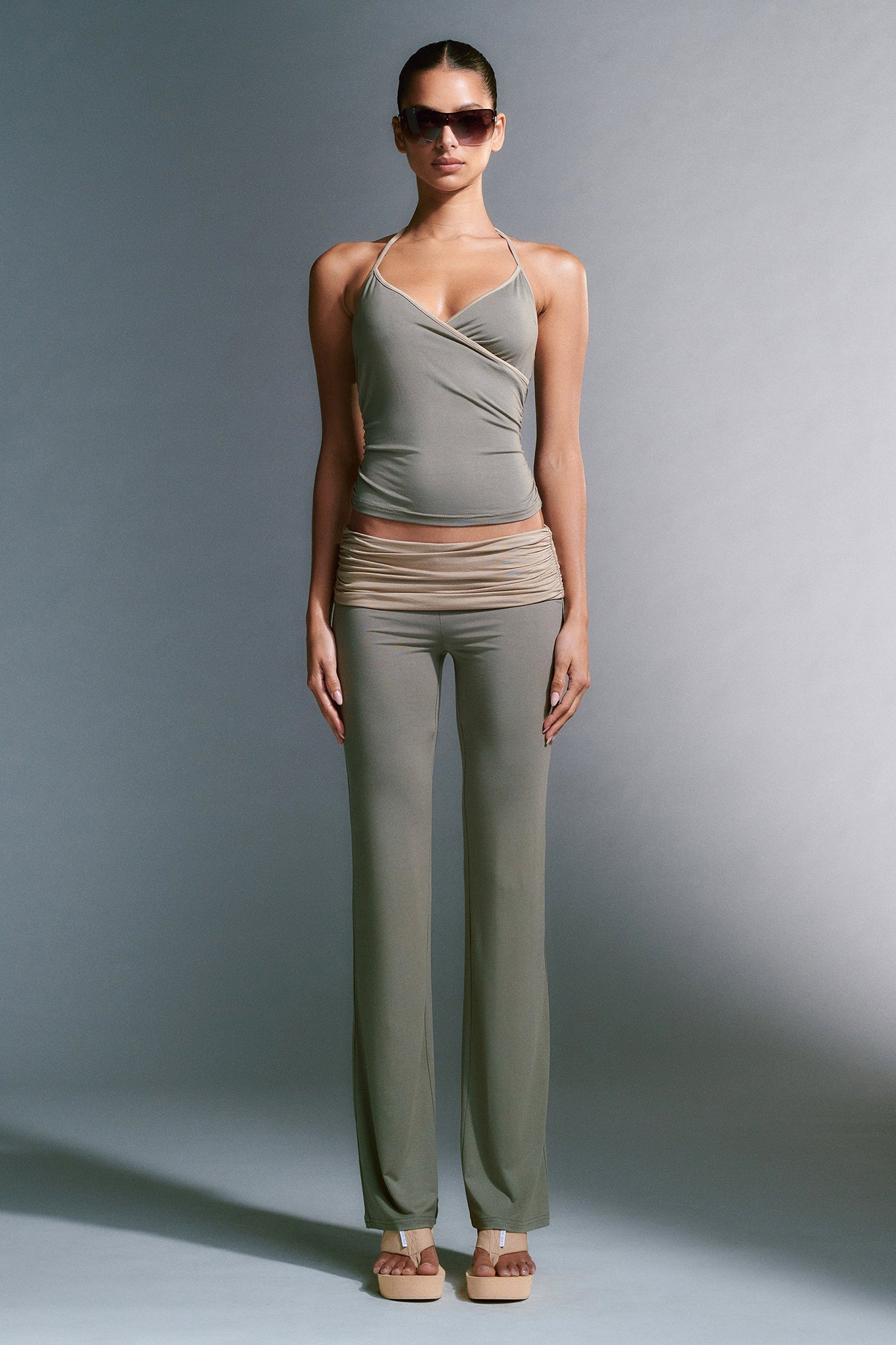 A person stands against a gray background, showcasing the JANNIS TANK - GREEN from I.AM.GIA and matching wide-leg pants. With perfect posture and hands at their sides, they sport dark sunglasses and beige sandals. The stylish outfit features a wrap style on the top and elegant ruching.