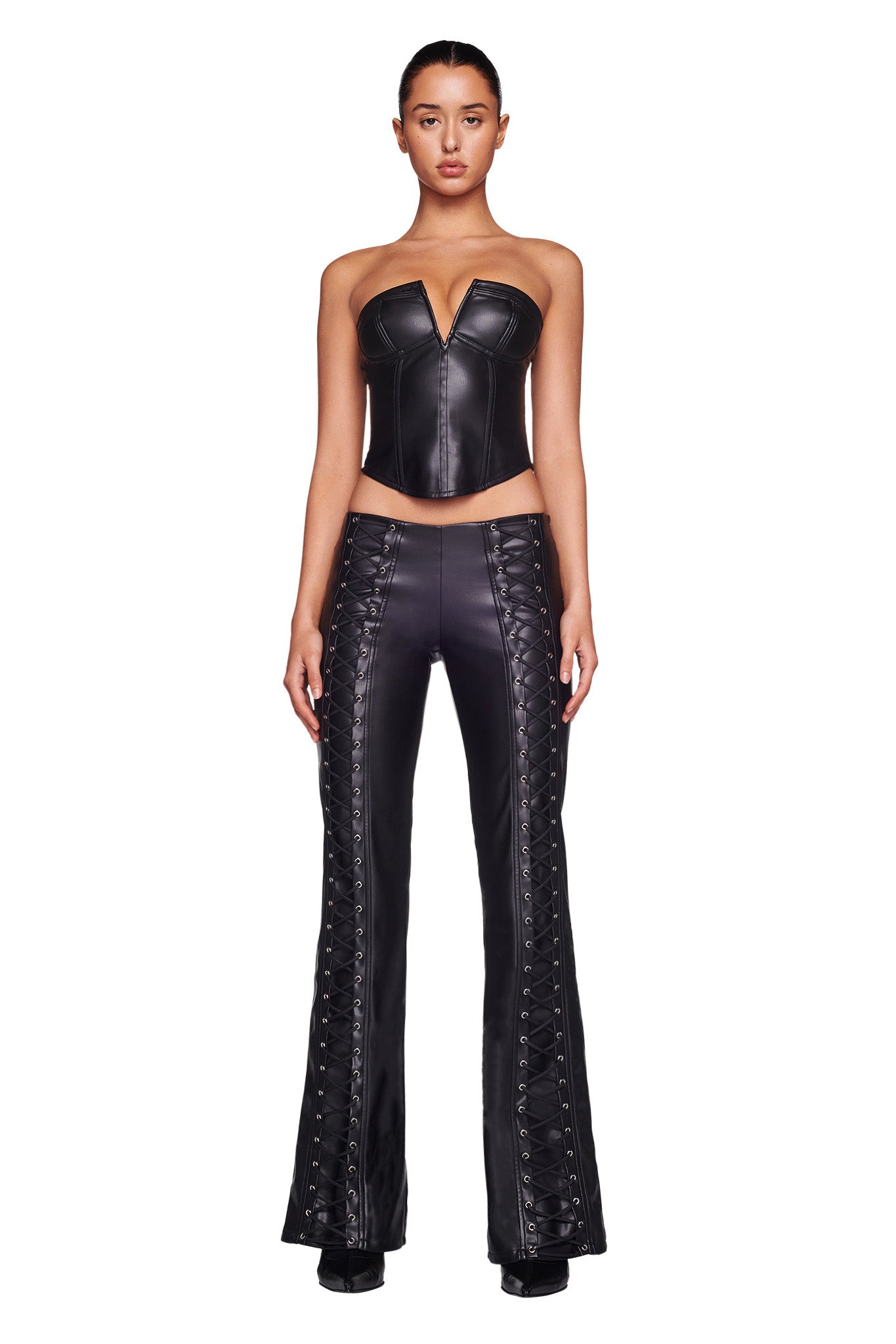 A woman is standing against a white background, wearing the EVANNA CORSET - BLACK by I.AM.GIA, a black, strapless, form-fitting leather top with a plunging neckline. She pairs it with matching high-waisted leather pants featuring lace-up detailing on the sides. She stands confidently, facing forward.