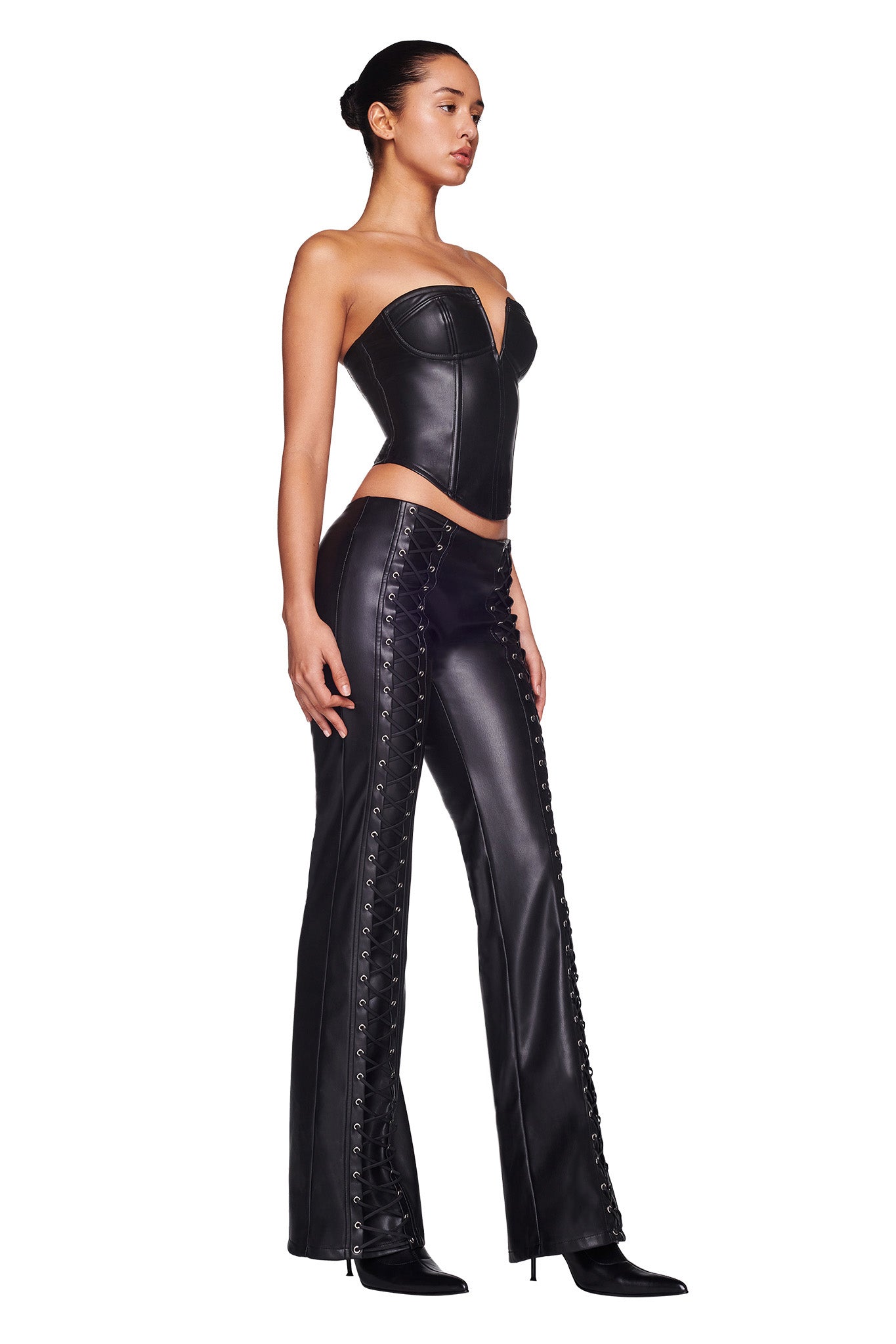 A woman stands facing left, wearing the EVANNA CORSET - BLACK from I.AM.GIA and matching I.AM.GIA black leather pants featuring side lace-up details. Her hair is tied back in a bun, and she pairs the outfit with black high-heeled boots. The background is plain white.