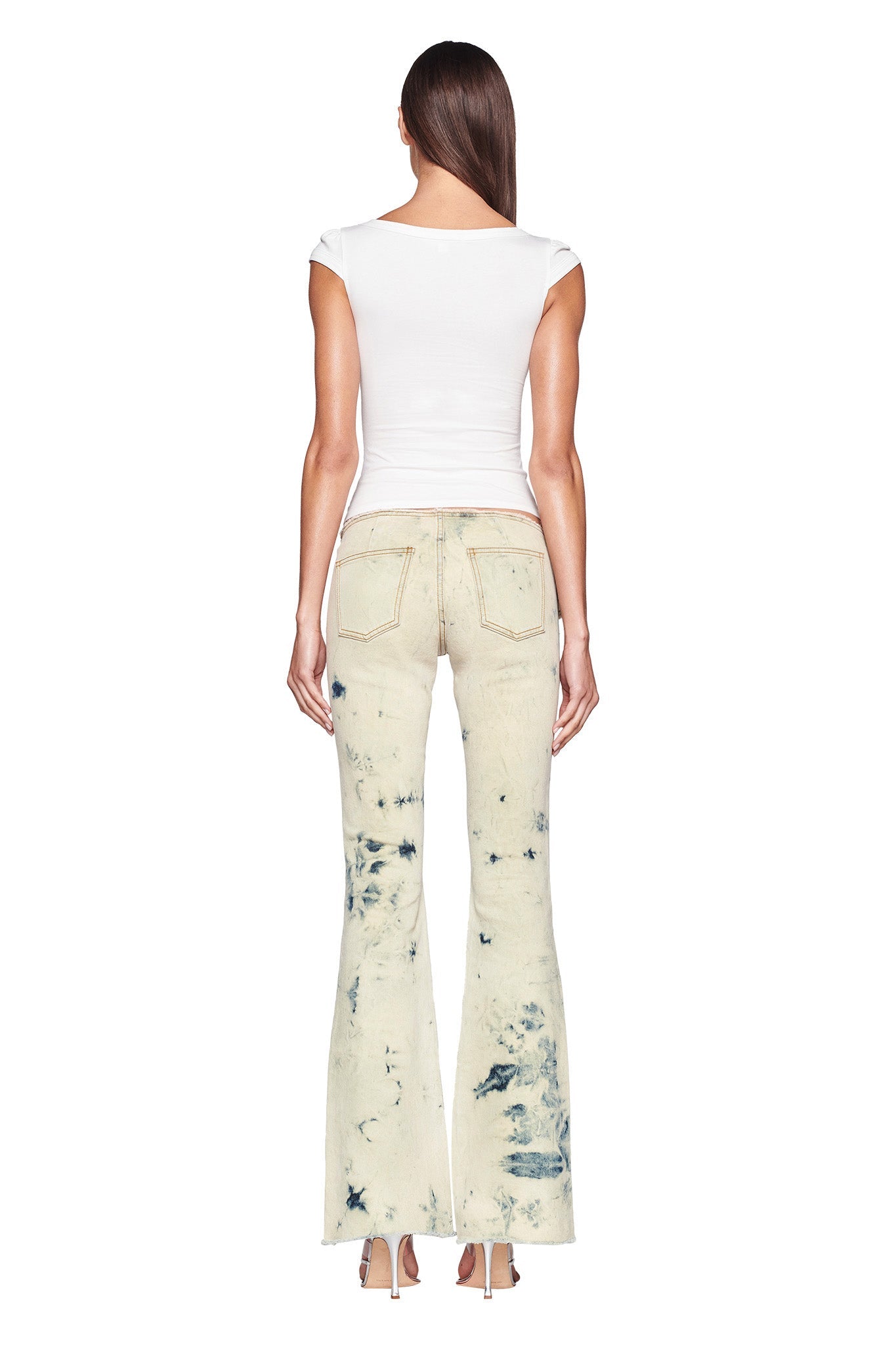 A person stands facing away, dressed in the white GIULIA TEE from I.AM.GIA and high-waisted, flared I.AM.GIA jeans featuring a bleached, distressed pattern. The individual has long, straight brown hair and is wearing high-heeled sandals. The background is plain white.
