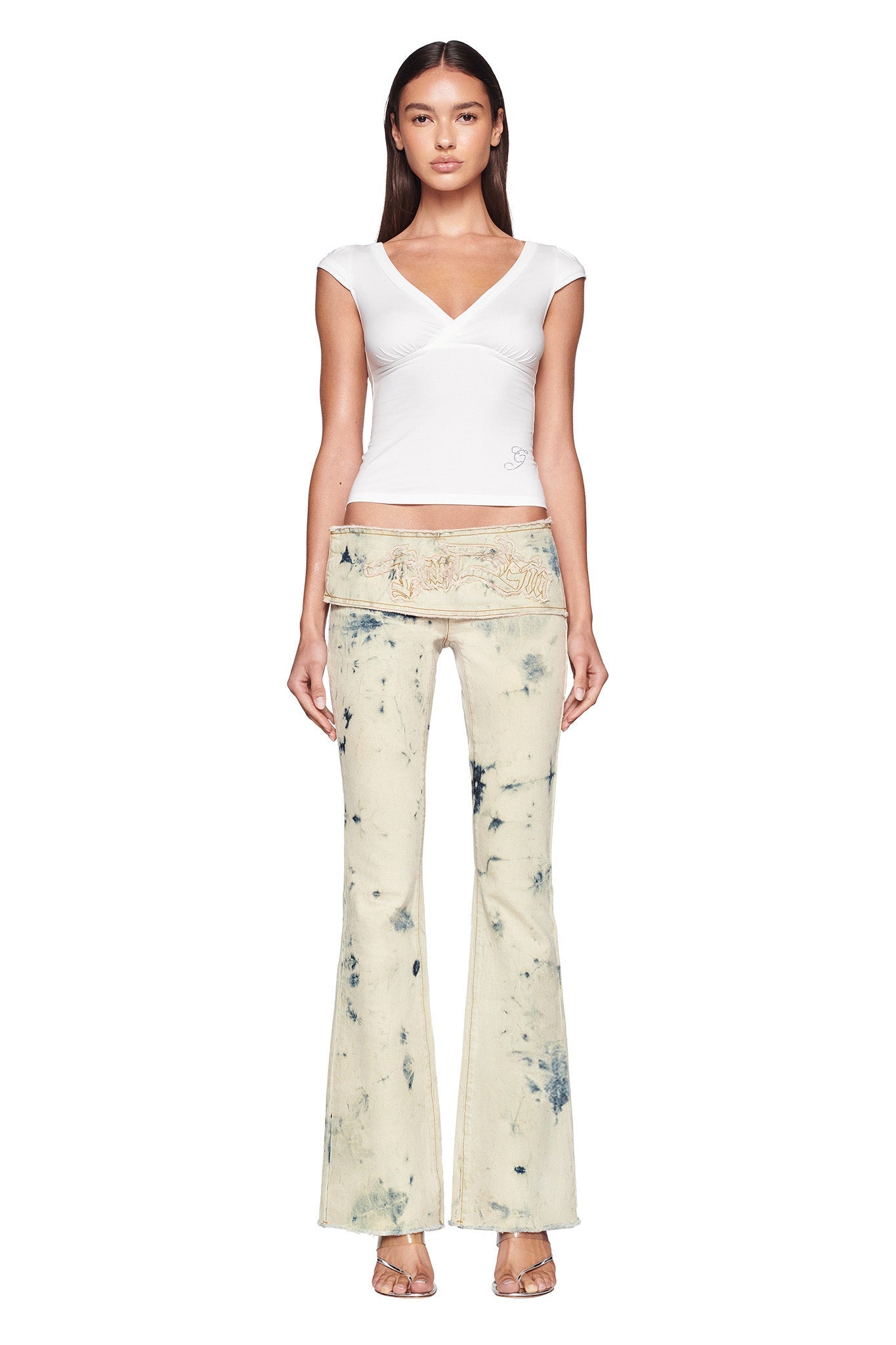 A woman with long hair stands facing the camera, wearing a fitted white GIULIA TEE from I.AM.GIA and tie-dye beige pants by I.AM.GIA with a low-rise waist and flared legs. She also sports open-toe beige heels. The background is plain white.