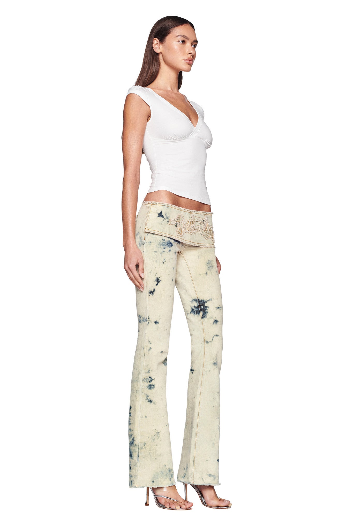 A woman stands in a side profile pose, showcasing her I.AM.GIA GIULIA TEE - WHITE paired with acid-washed light blue high-waisted jeans featuring an embroidered floral pattern on the waistband. Her high-heeled sandals complete the look, and her straight hair is worn down.