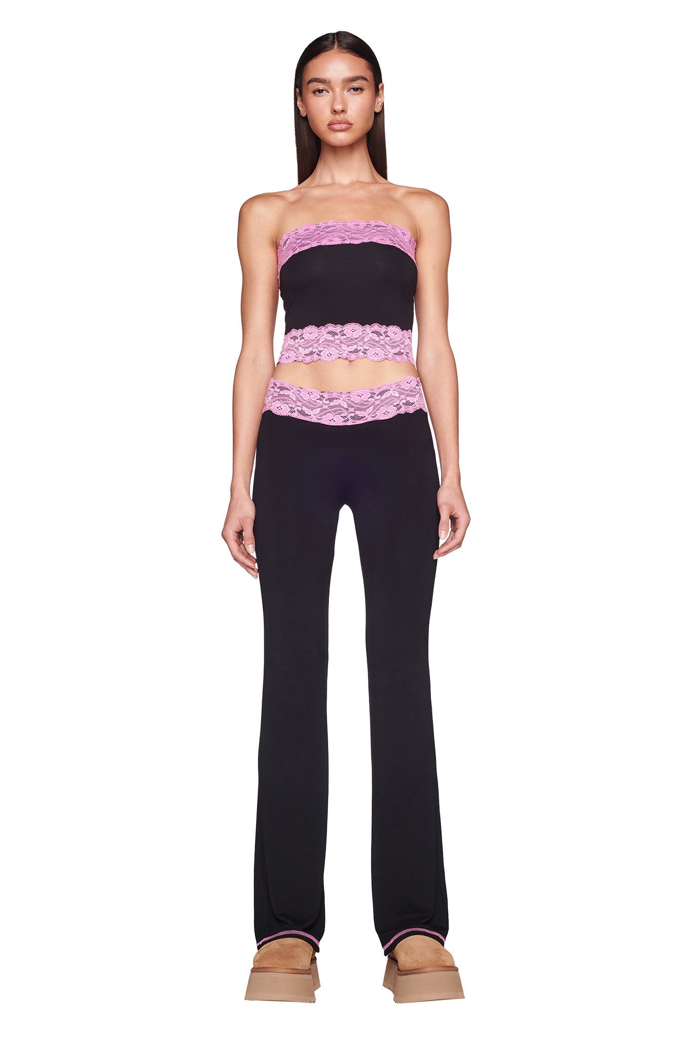 A woman is standing against a plain white background, showcasing an I.AM.GIA black strapless top with pink lace trim and the TARSHA PANT in black/pink, featuring matching pink lace at the waist. She completes her look with beige platform shoes. Her long dark hair is straight and she has a serious expression.