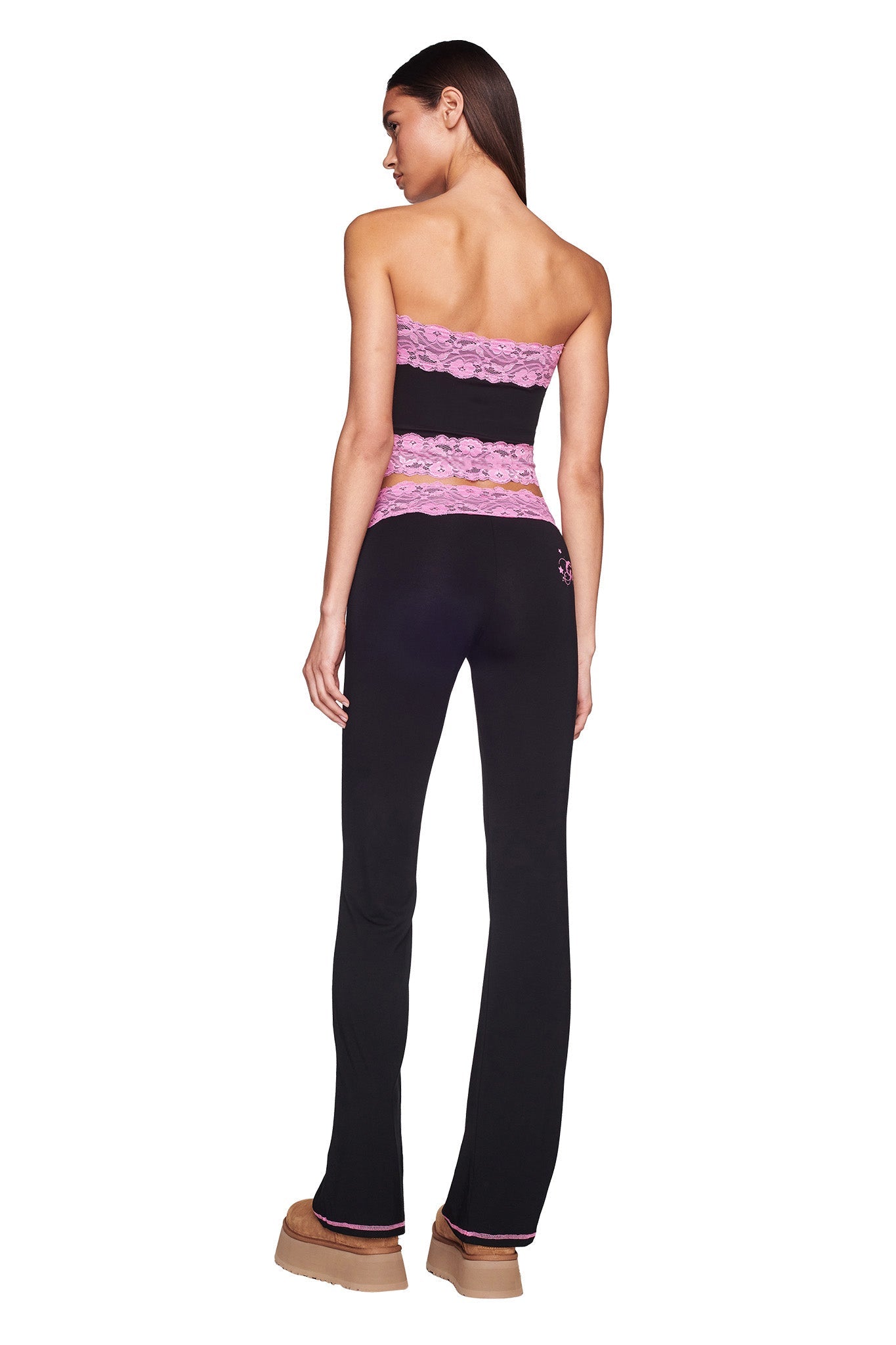 A woman with long brown hair stands with her back to the camera, wearing the TARSHA PANT - BLACK/PINK from I.AM.GIA, featuring strapless design and pink lace detailing around the bust and waist. She pairs the outfit with tan platform shoes.