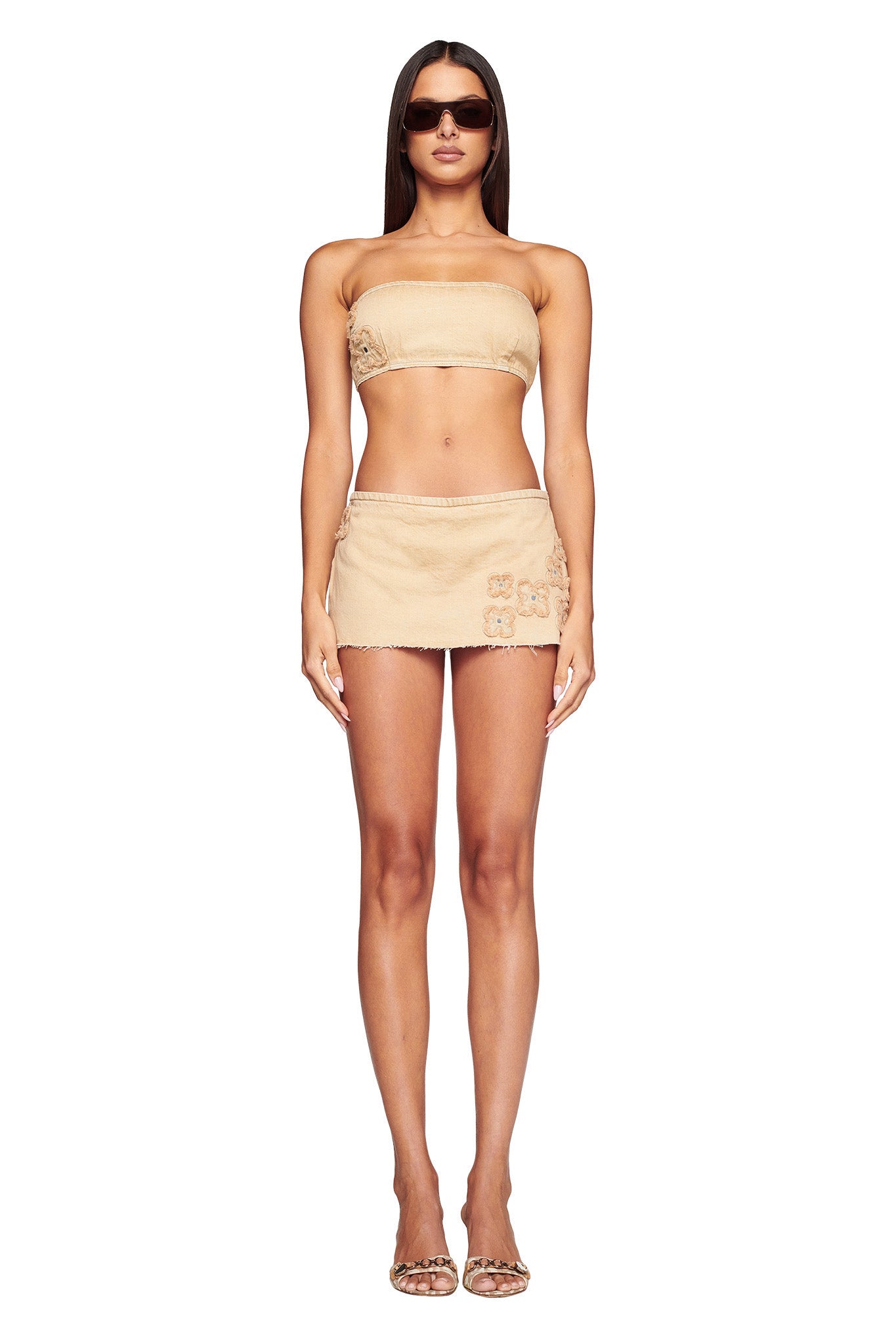 A woman stands facing forward, wearing a tan strapless bandeau top paired with the FAUNA MINI SKORT - BEIGE in tan denim by I.AM.GIA, which features flower embellishments on the side. She has long, straight hair and is accessorized with dark sunglasses and clear high-heeled sandals.