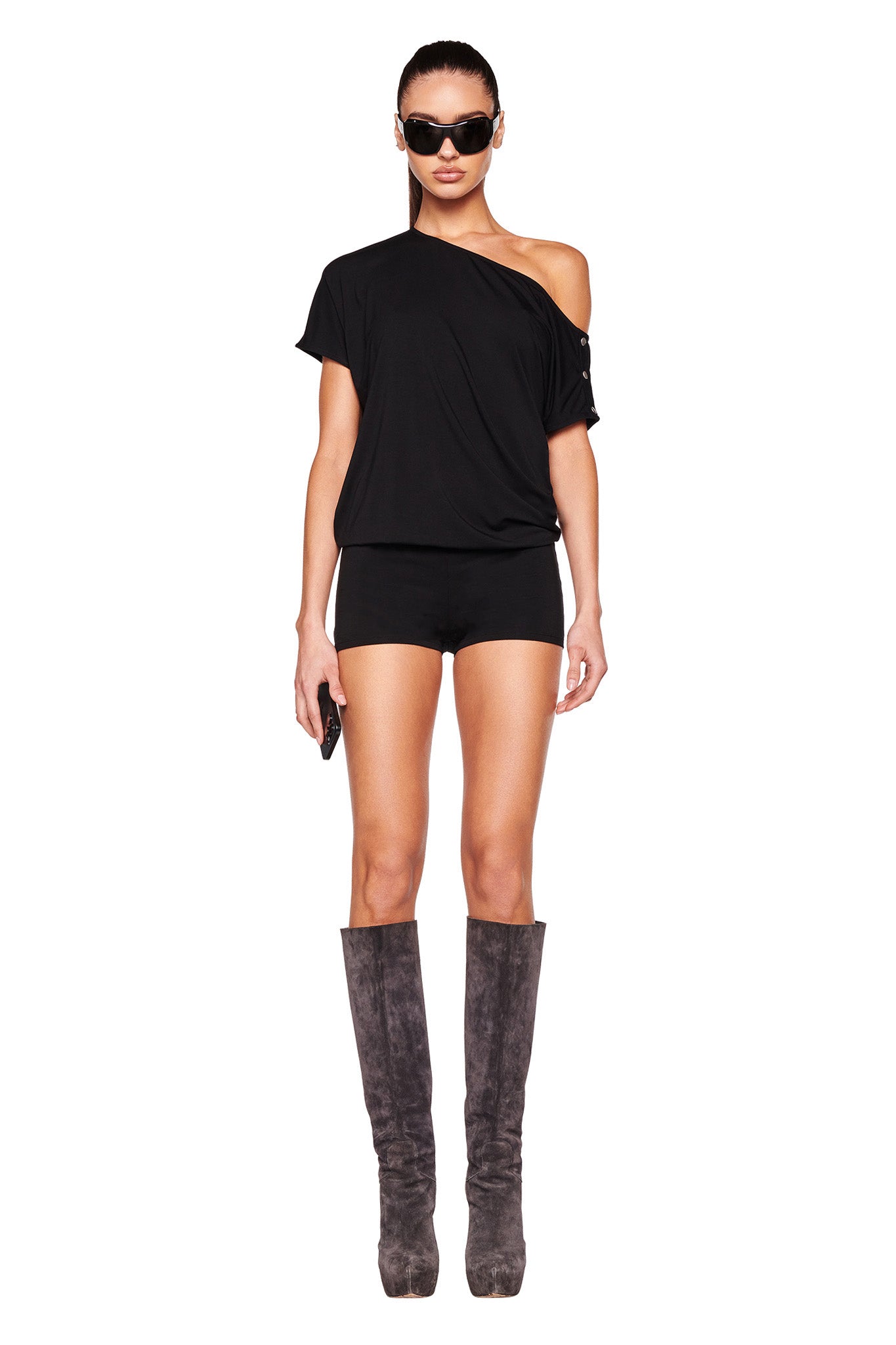 A person stands against a white background wearing the black I.AM.GIA EZIA ROMPER and tall grey boots. They are also wearing dark sunglasses, holding a small object in their hand. The outfit channels that edgy I.AM.GIA aesthetic perfectly.