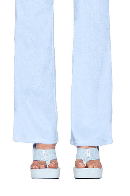 A close-up photo features the lower half of a person wearing light blue wide-leg pants by I.AM.GIA and matching SHELBY FLIP FLOP - BLUE : BABY BLUE sandals from the same brand. The flip flops have thick straps over the toes and ankles, perfectly complementing the ensemble. The person's toenails are painted a soft, light pink.