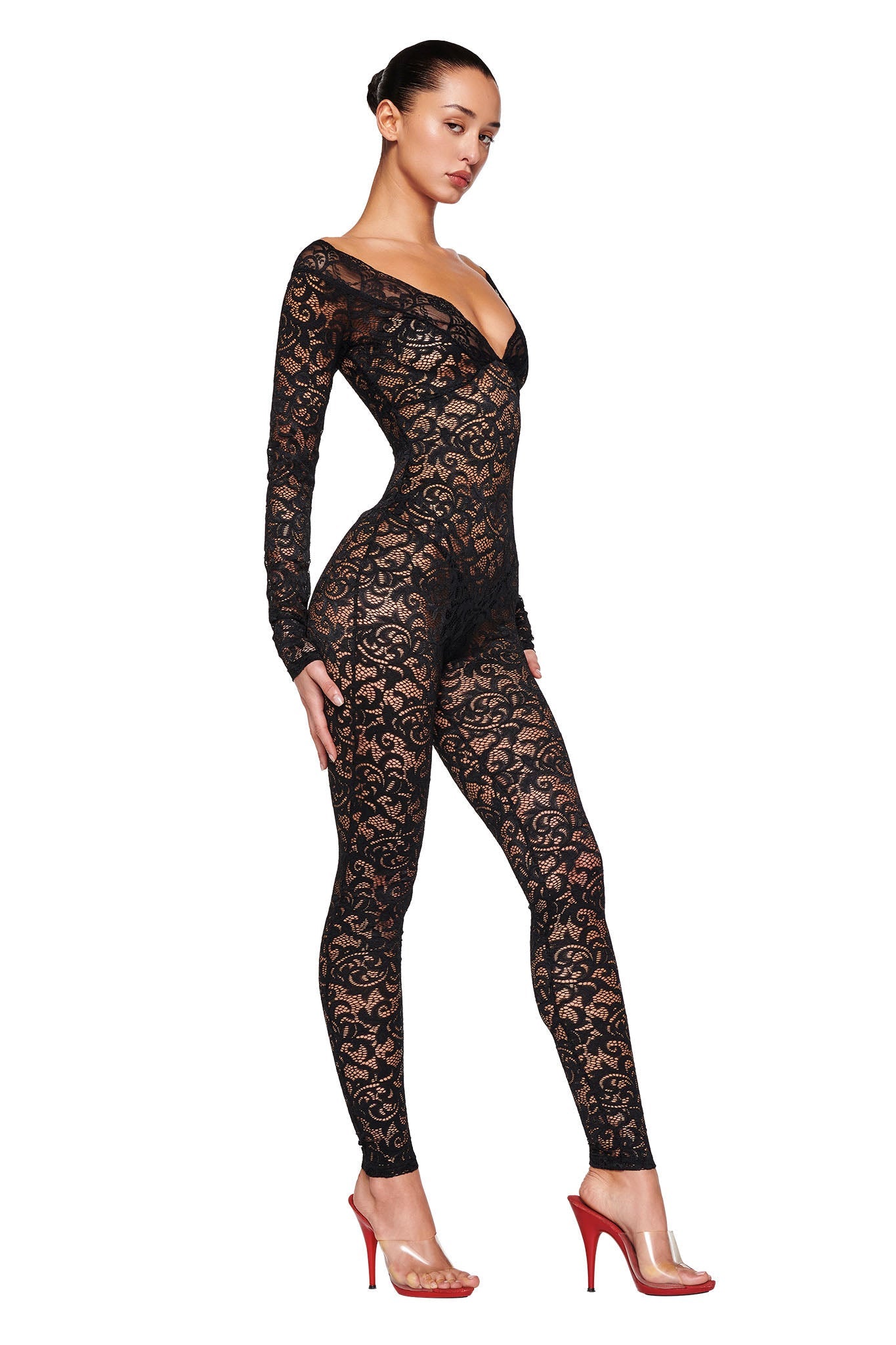 A person stands confidently, modeling the GEMMA CATSUIT - BLACK by I.AM.GIA. They are wearing clear high-heeled shoes with red soles. The person is posing with one leg slightly forward and looking off to the side, against a plain white background.