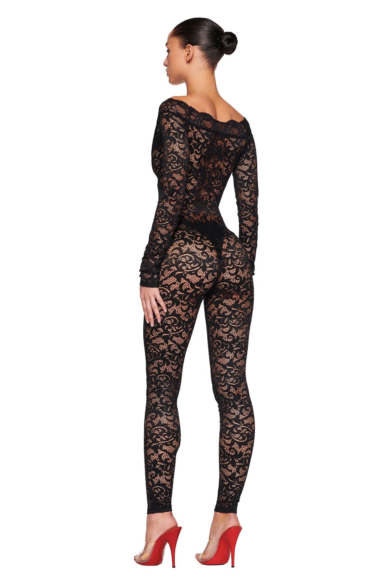 A woman stands with one leg slightly bent, wearing the GEMMA CATSUIT - BLACK by I.AM.GIA, a semi-transparent black lace jumpsuit featuring a floral pattern. She pairs it with red high heels. Her hair is styled in a neat bun, and she faces away from the camera, showcasing the back of her stunning outfit.
