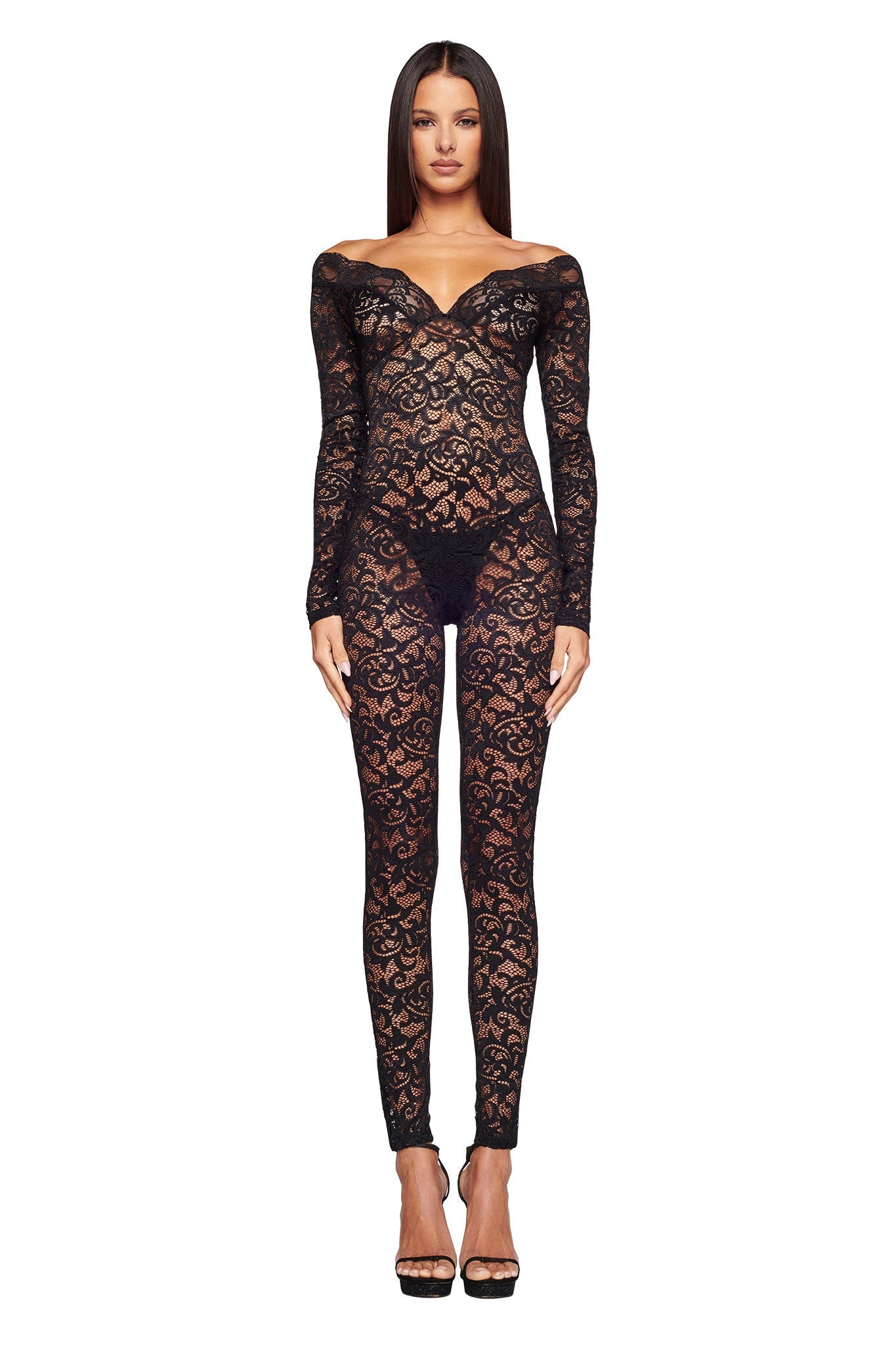 A person stands facing forward, wearing the GEMMA CATSUIT - BLACK by I.AM.GIA. This off-the-shoulder, long-sleeve jumpsuit is made of sheer black lace and features an intricate floral pattern. They are also wearing black high-heeled shoes.