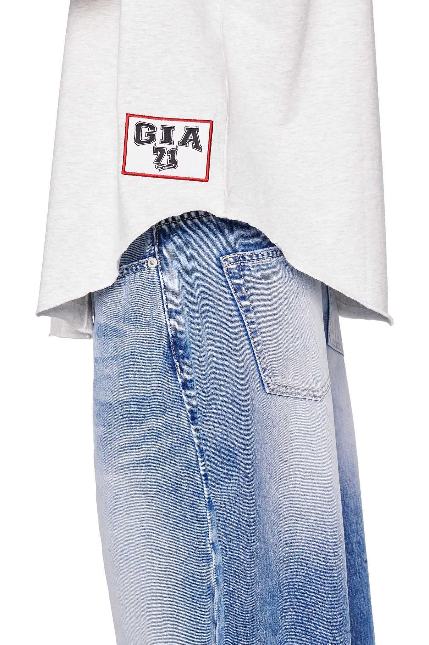 A close-up of a person wearing high-waisted, light-wash jeans and the "LORRI JERSEY - GREY" from I.AM.GIA, featuring an asymmetrical hem. The top has a patch near the bottom with the text "GIA 23" in black and red, subtly showcasing the I.AM.GIA style. The image focuses on the lower back portion of the outfit.
