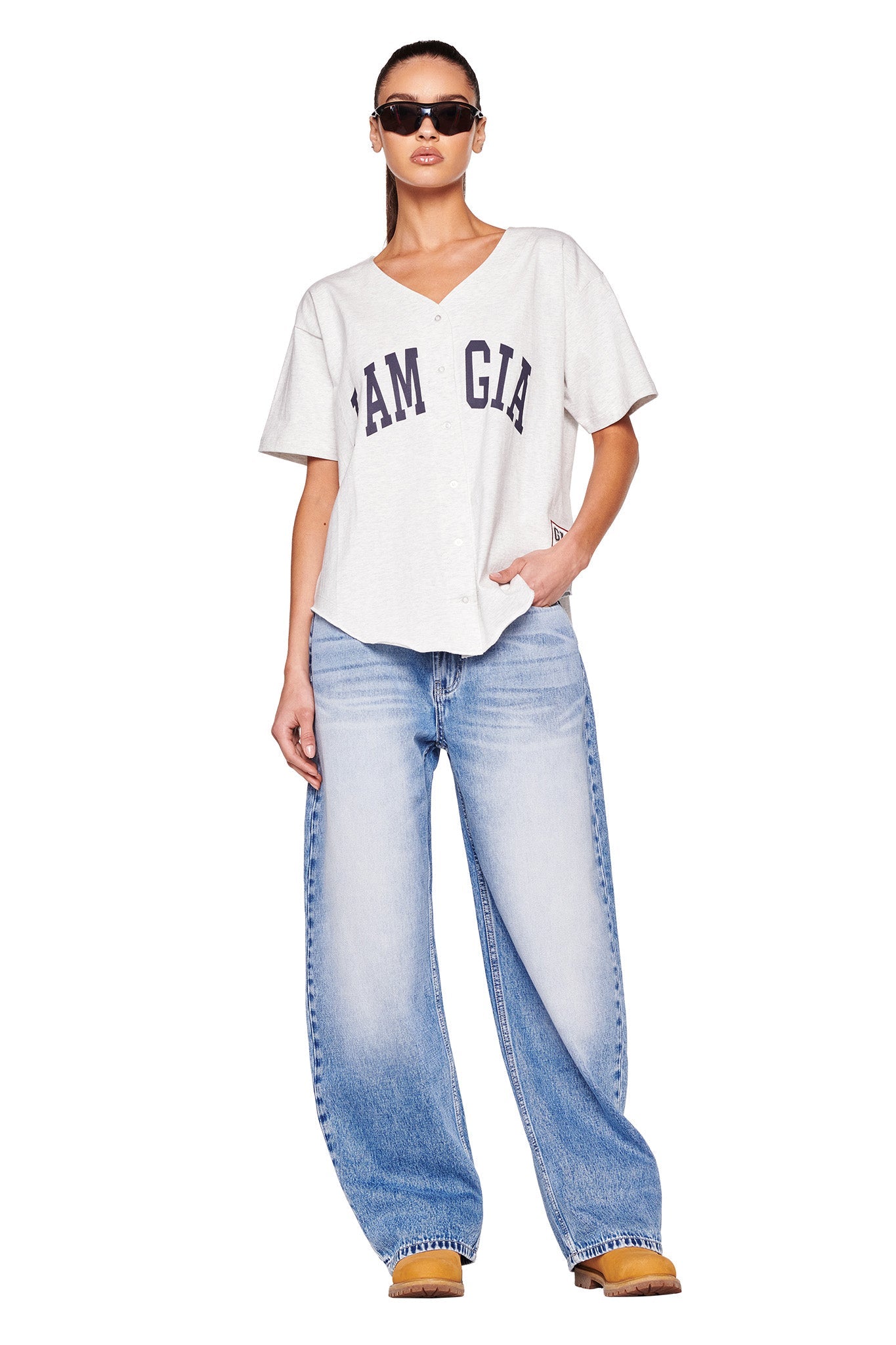 A person with long hair stands wearing sunglasses. They are dressed in a light gray LORRI JERSEY from I.AM.GIA, loose light blue jeans, and tan shoes. One hand is in their pocket, and they have a relaxed posture. The background is white.