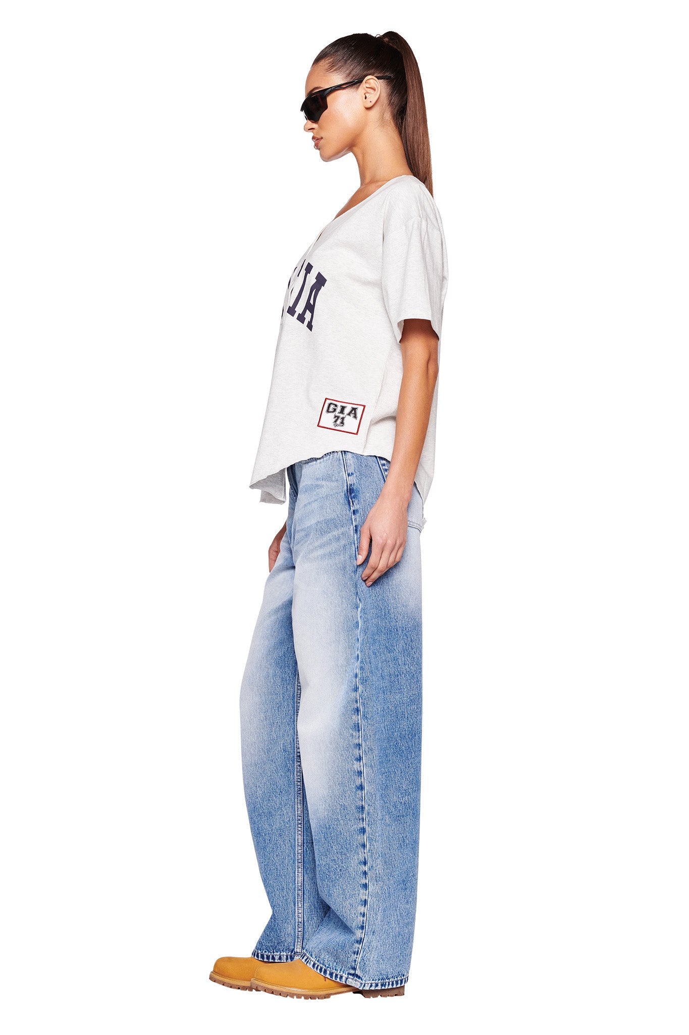 A person with long hair tied back in a ponytail, wearing sunglasses, a loose-fitting light grey LORRI JERSEY by I.AM.GIA with text on it, and baggy light blue jeans. They are also wearing orange shoes and standing in a side profile position against a plain white background.