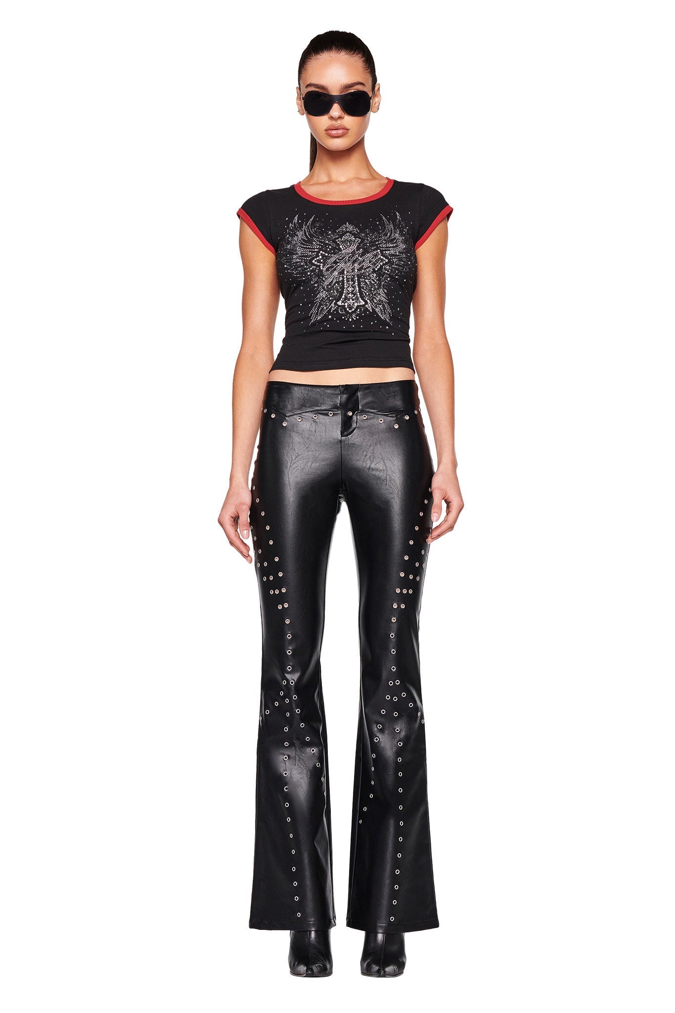 A person stands facing forward, wearing black sunglasses and the fitted DORA TOP - BLACK by I.AM.GIA, featuring a graphic design. They pair it with shiny black leather pants adorned with silver eyelets. Against a white background, they exude a bold and confident look.