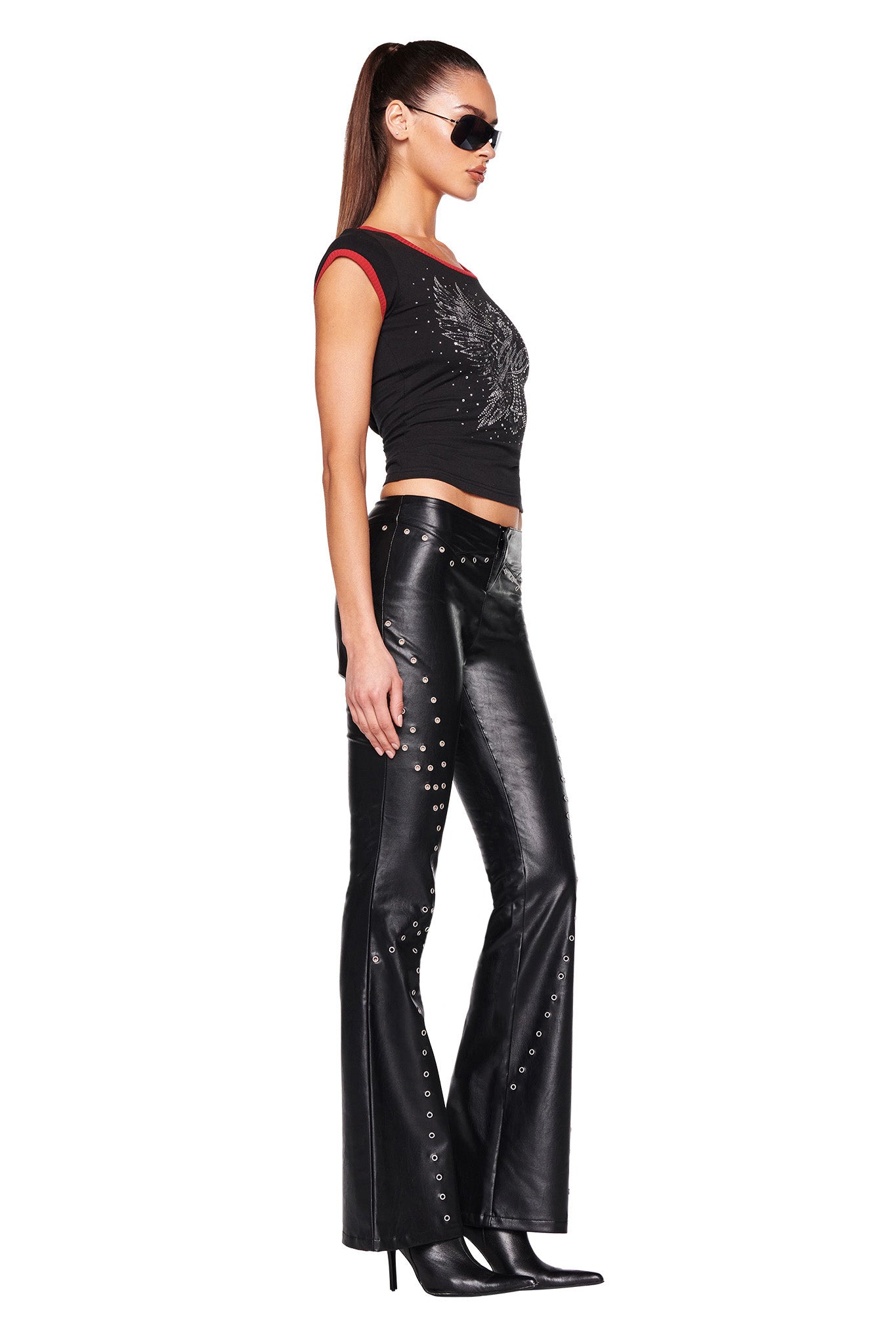 A woman stands in a side profile wearing I.AM.GIA sunglasses, the DORA TOP - BLACK sleeveless top with red trim from I.AM.GIA, and black leather pants featuring metallic studs. She completes the look with black heeled boots.
