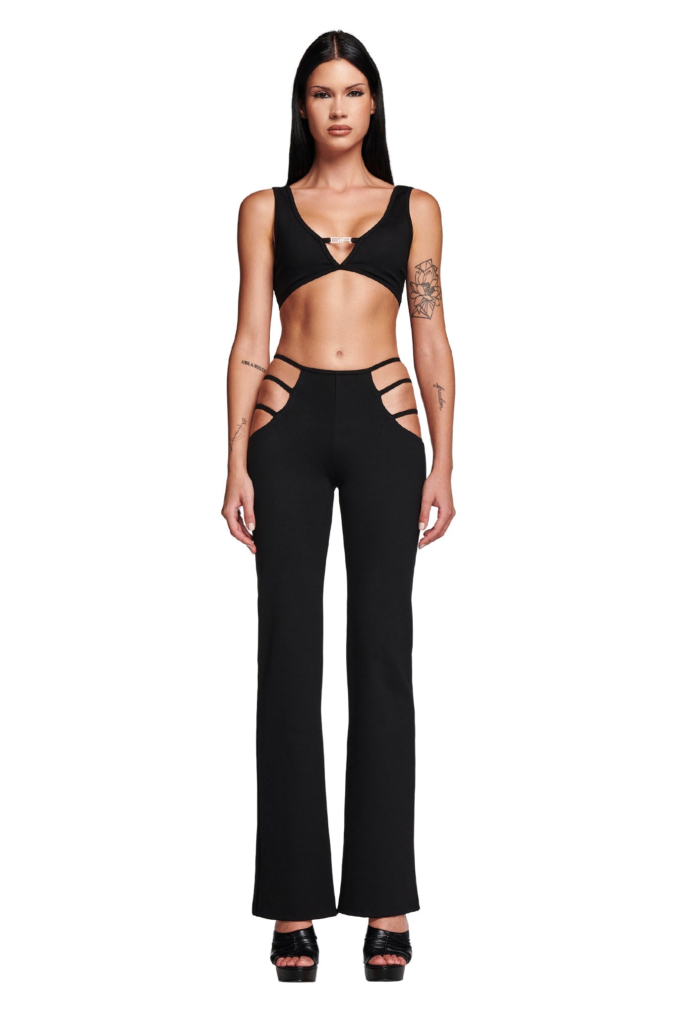 A woman with long, dark hair stands against a white backdrop. She is wearing the black LUCID TOP from I.AM.GIA and a pair of high-waisted pants with cut-out details on the hips. Visible tattoos adorn her right upper arm and left forearm, complementing her black platform sandals.