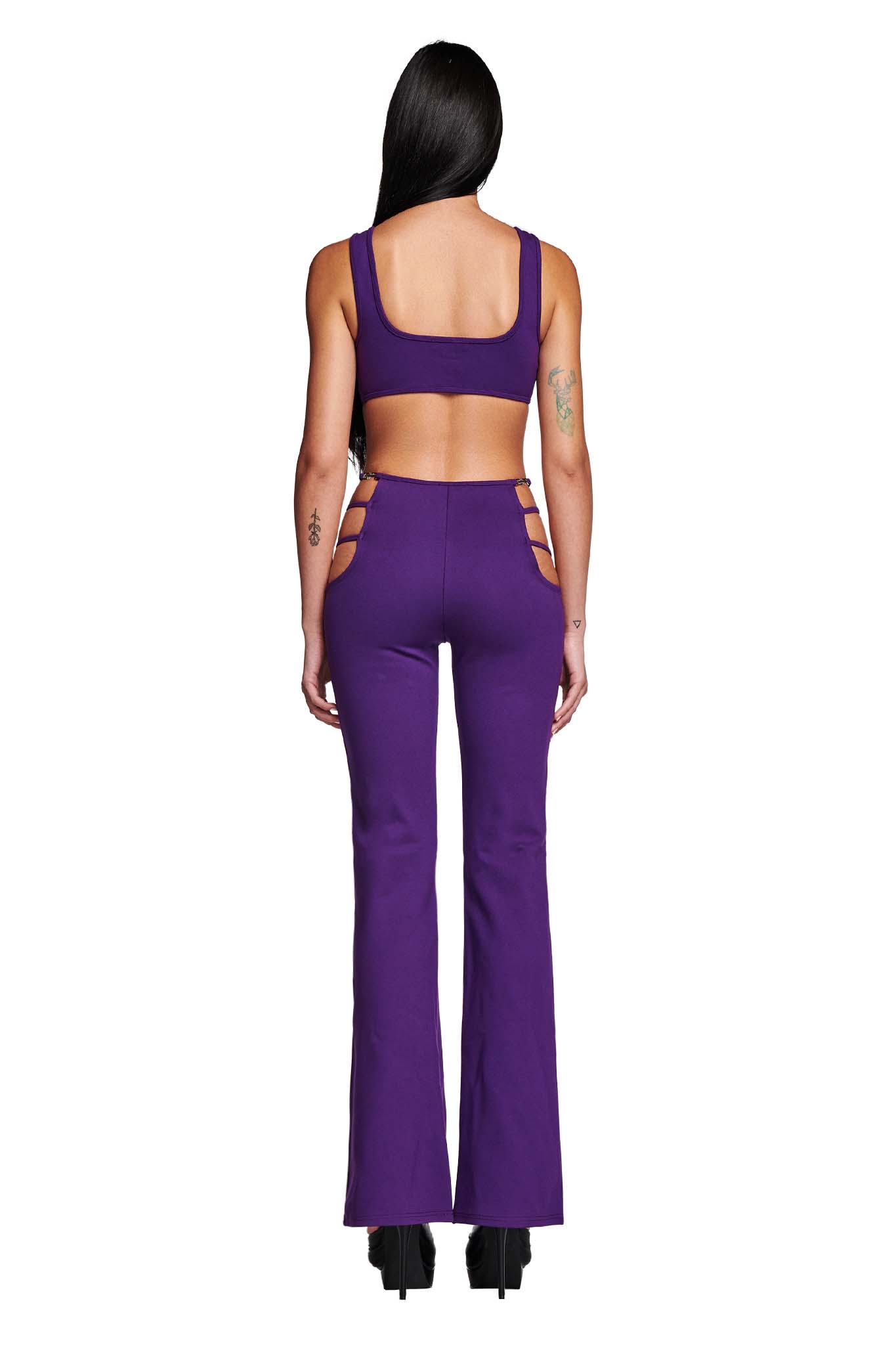 A person with long dark hair is standing with their back to the camera, showcasing the LUCID TOP - PURPLE by I.AM.GIA. The matching ensemble features a crop top and flared pants with side cutouts that reveal tattoos on their arms. They complete the edgy look with black platform shoes.