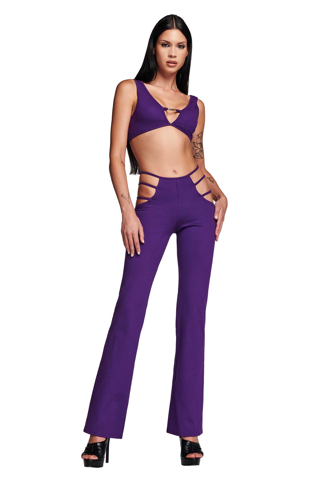 A woman is confidently modeling a bold I.AM.GIA outfit consisting of the LUCID TOP - PURPLE, a crop top with cut-outs, and matching high-waisted pants with side cut-outs. She is wearing black high-heeled sandals, has long straight hair, and poses with one hand on her hip.